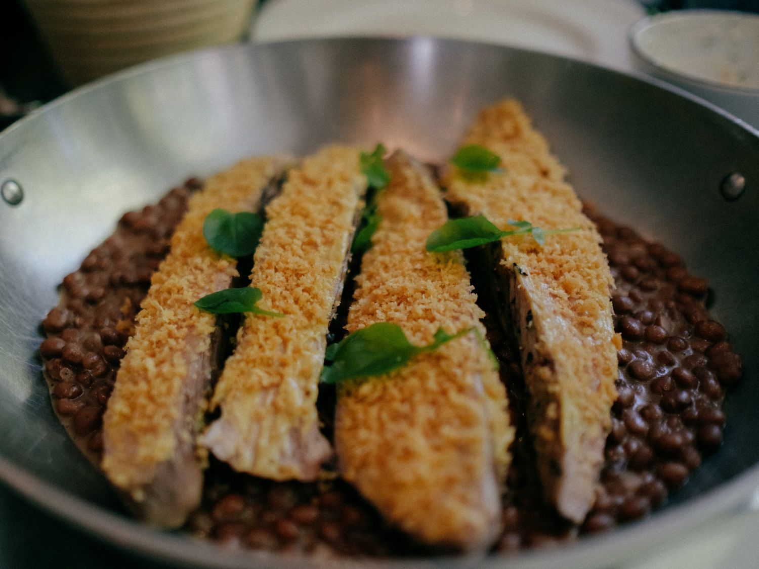 Crispy sesame-crusted fish fillets served over dark red beans in a silver pan, garnished with fresh herbs.