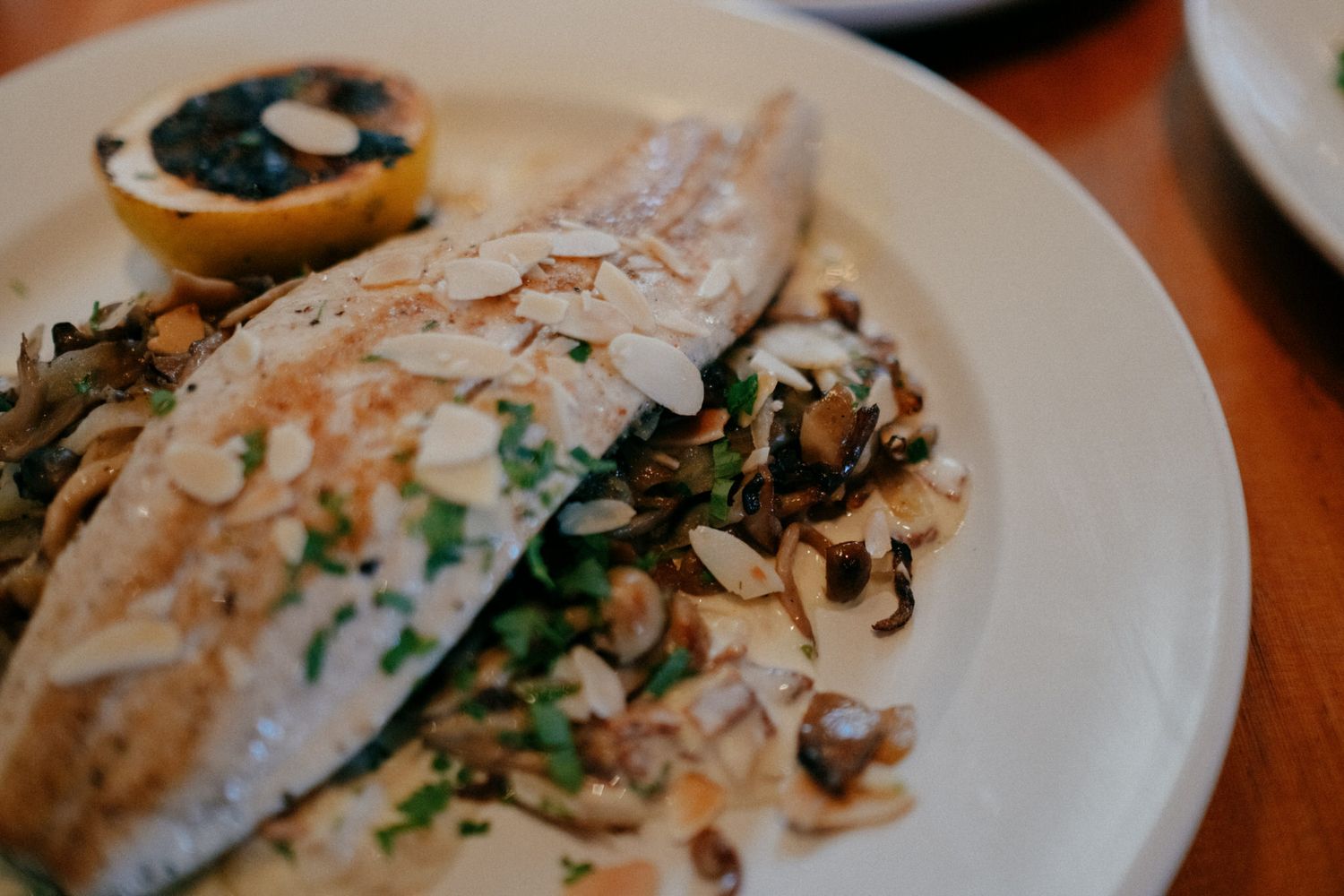 Grilled fish fillet topped with sliced almonds and chives, served with sautéed mushrooms and a roasted lemon half on a white plate.