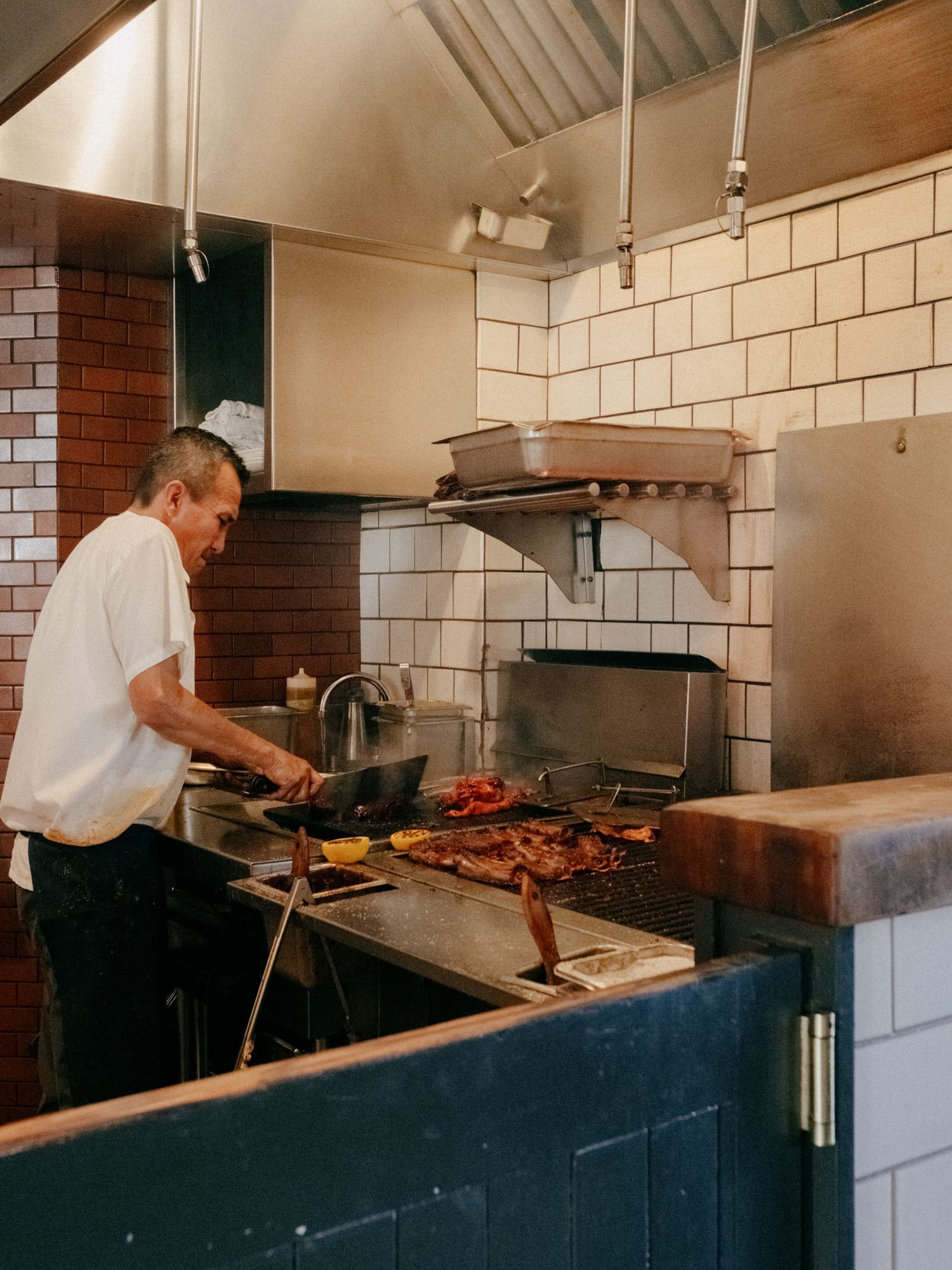 A chef grilling meat over an open flame in a rustic restaurant kitchen with white tiles and brick walls.