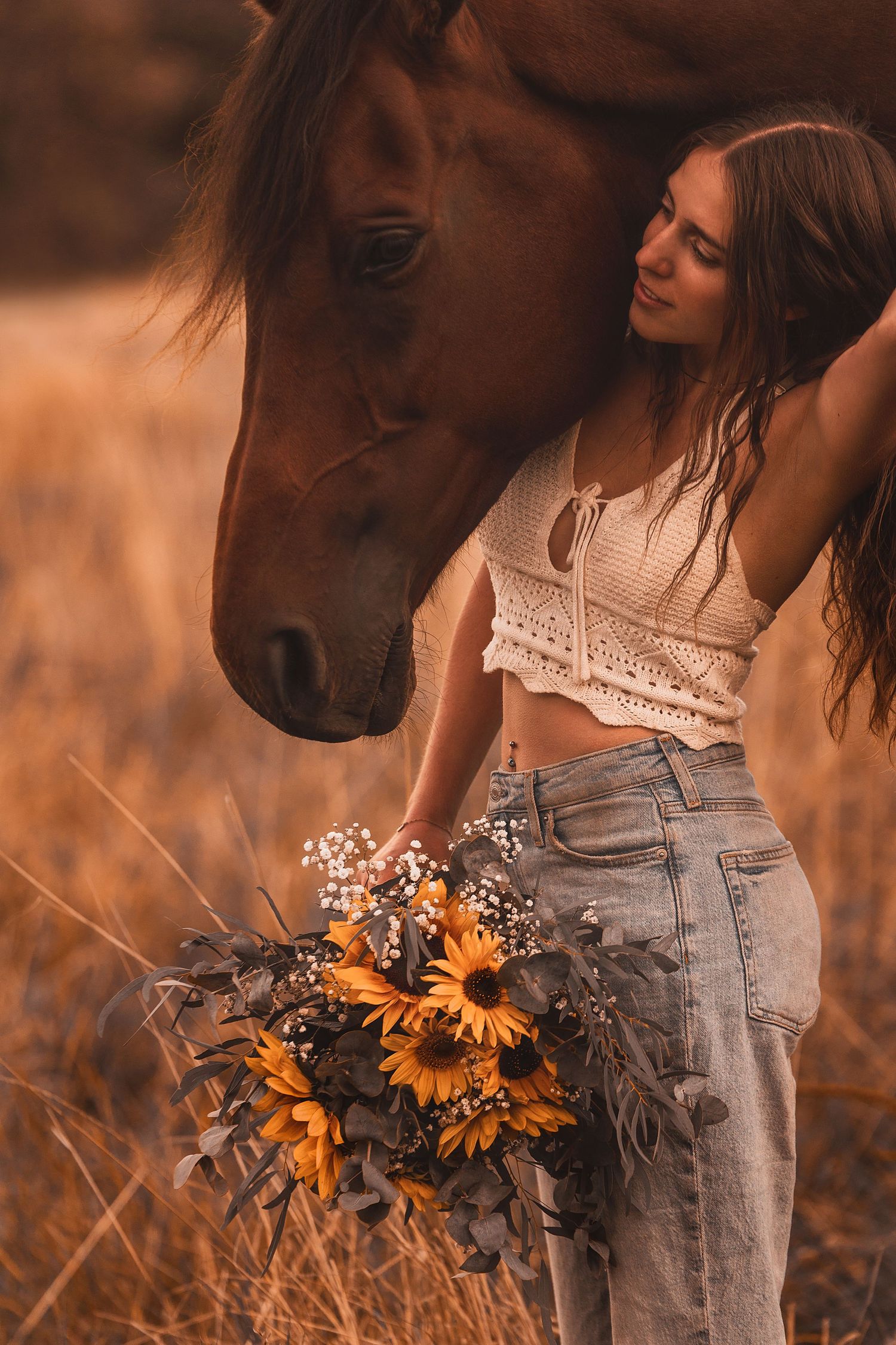A person in jeans holds a bouquet of sunflowers while standing next to a brown horse in a warm-toned field.