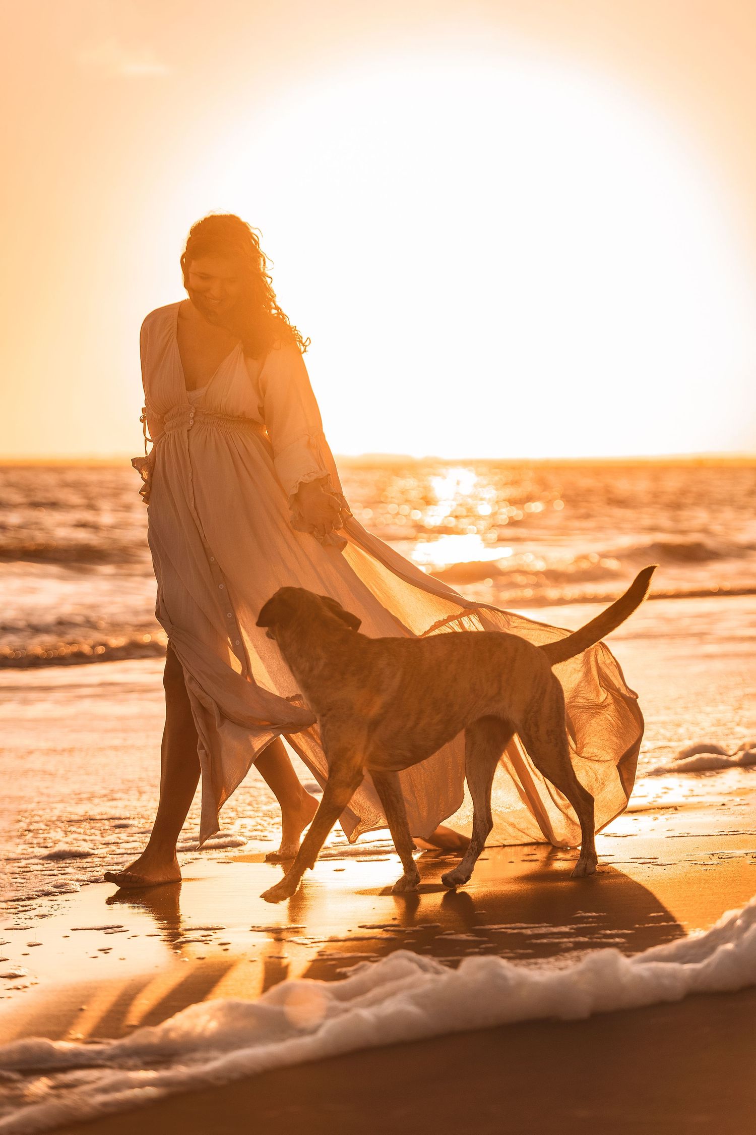 Frau im weißen Kleid geht bei goldenem Sonnenuntergang mit ihrem Hund am Strand entlang, während die Wellen ans Ufer plätschern.