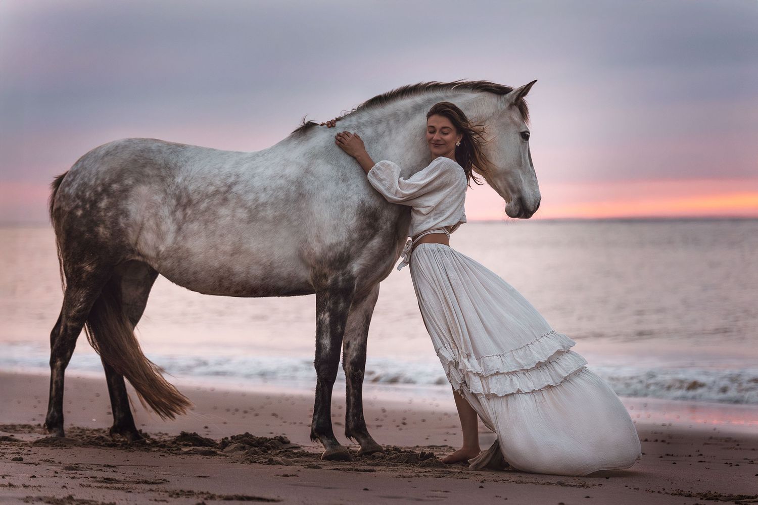 Eine Frau im weißen Kleid umarmt ein Schimmelpferd an einem Sandstrand bei Sonnenuntergang.