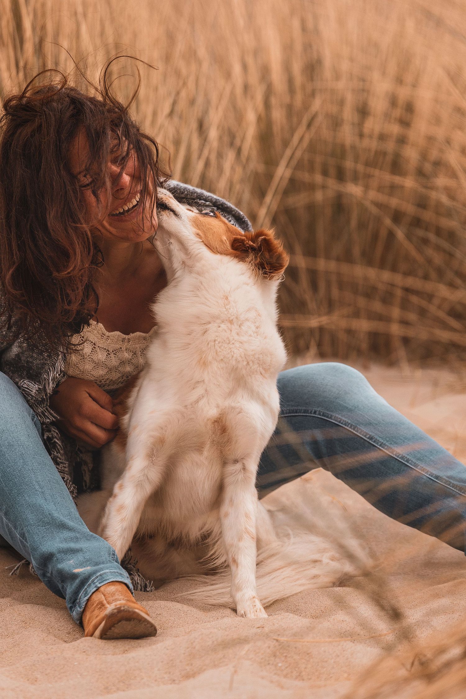 Eine lachende Frau mit lockigem Haar sitzt an einem Sandstrand, während ihr kleiner weißer Hund ihr liebevoll das Gesicht leckt.