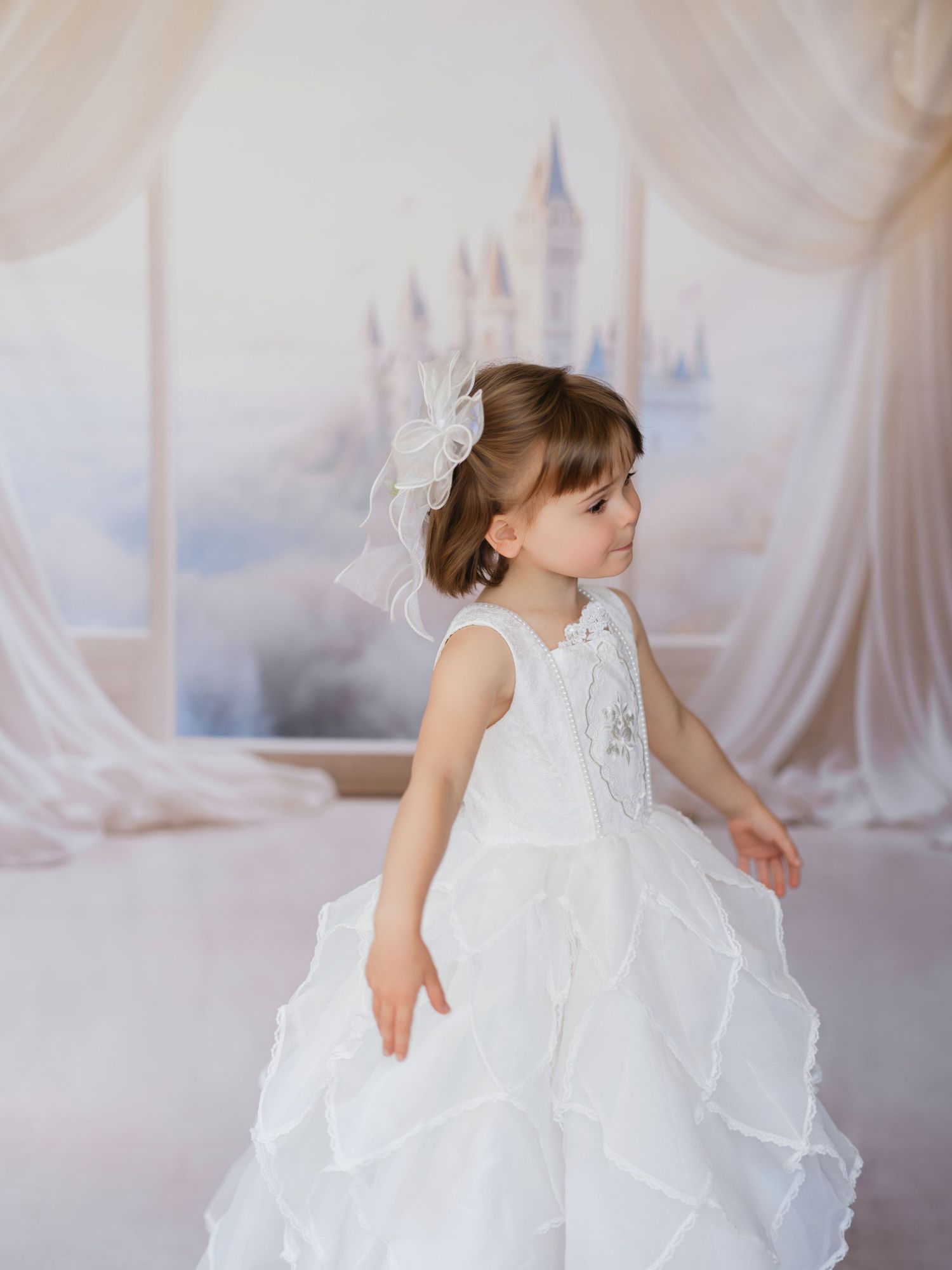 A young child in a flowing white lace dress twirls gracefully in a dreamy, light-filled studio setting.