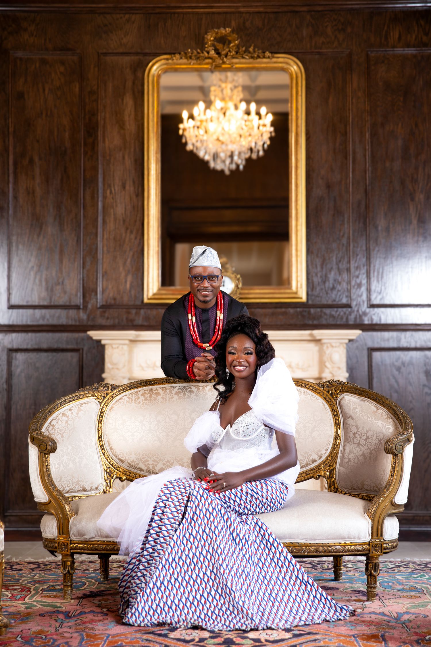 Elegant couple in traditional African attire pose on ornate gold and white settee beneath crystal chandelier and gilded mirror.