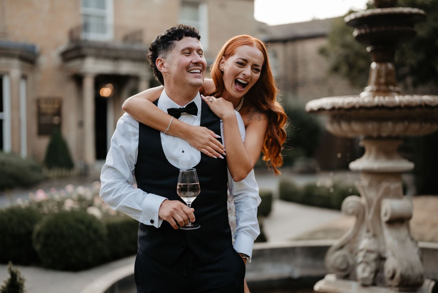 Happy couple laughing by a stone fountain, man in tuxedo vest holding wine glass, woman in white dress with red hair.