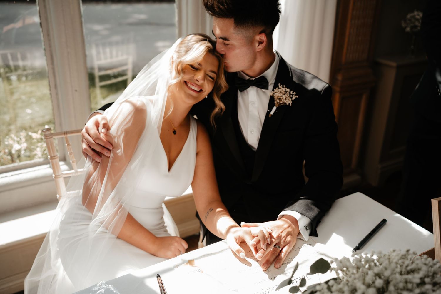 Bride and groom signing wedding register, sharing a tender kiss at a sunlit table with floral decor.