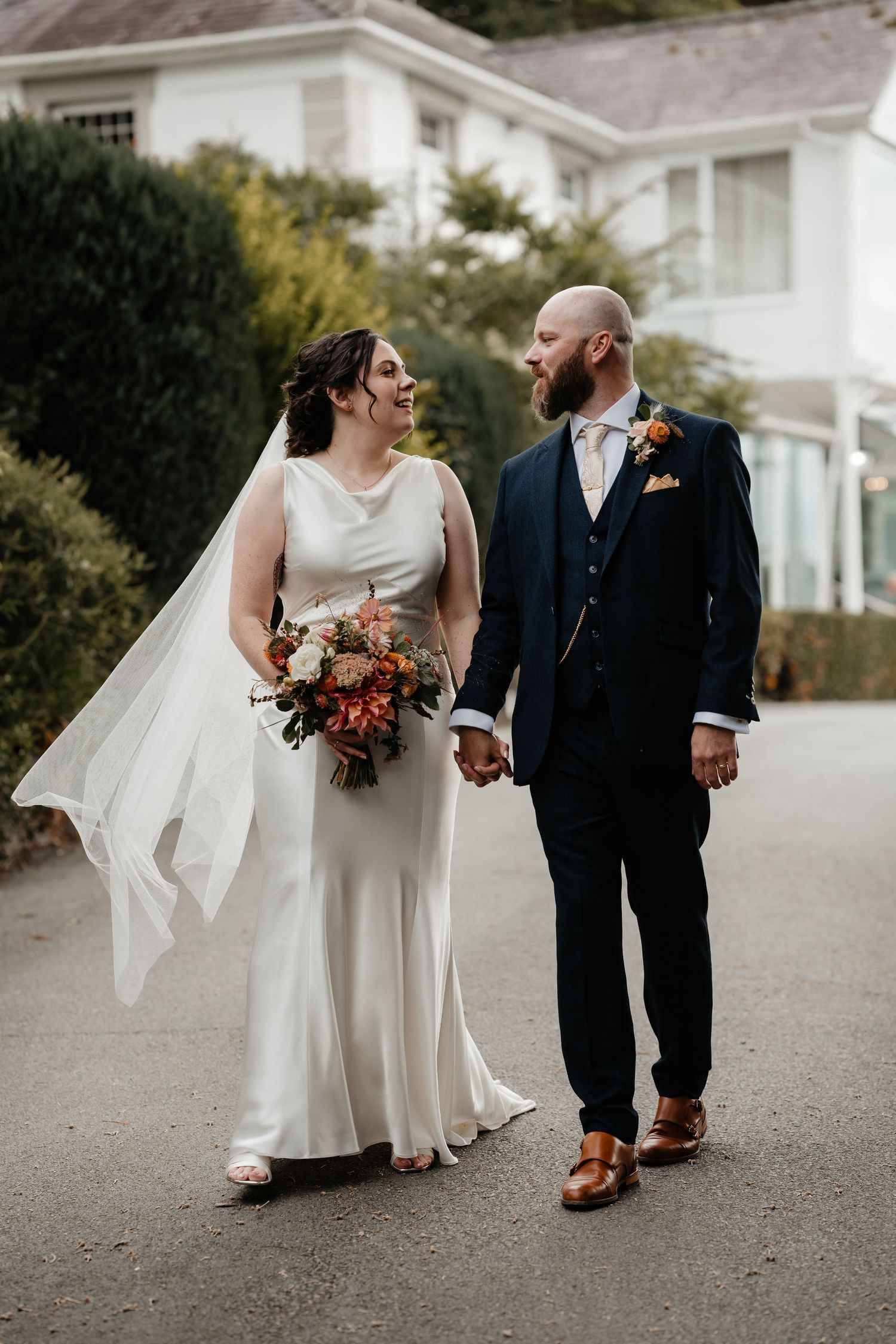 A bride and groom walk hand-in-hand outside a country house, she holds an autumn bouquet, he wears a navy suit.