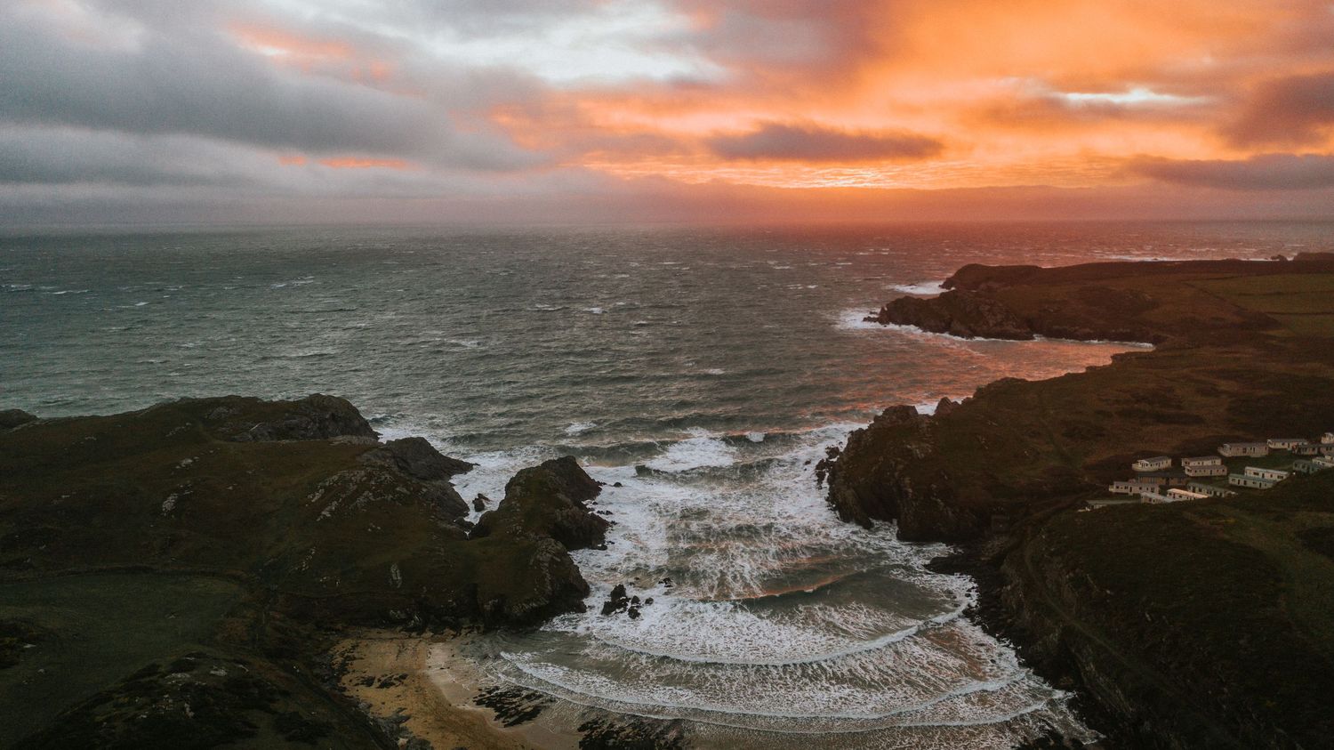 Aerial view of a dramatic coastal sunset with orange clouds over rocky shores and crashing waves.