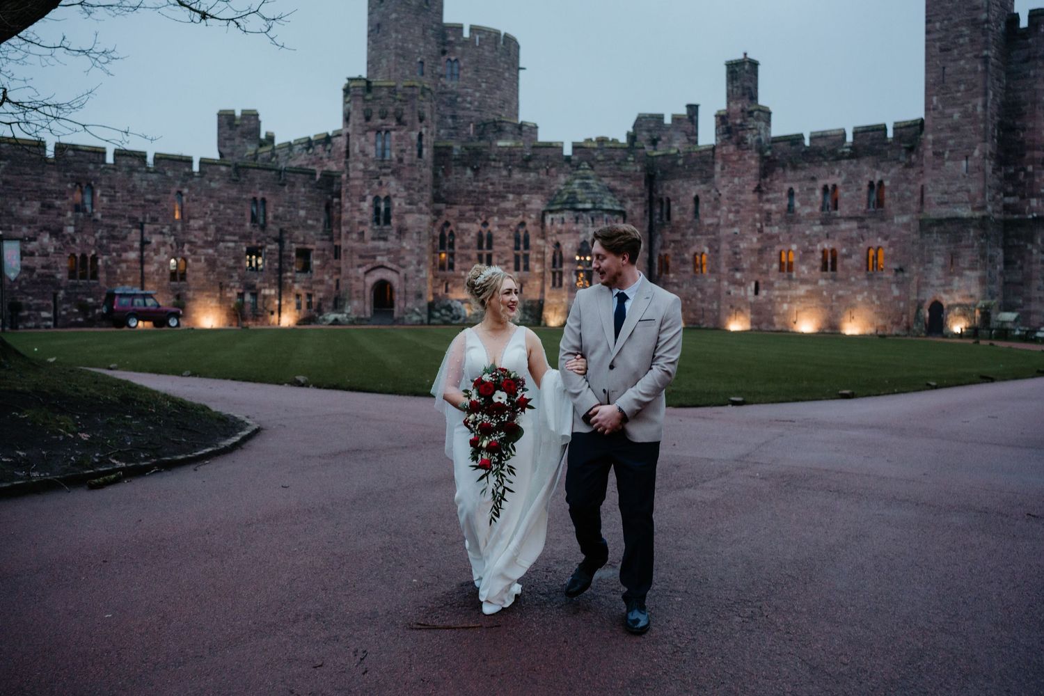 Bride and groom walk hand-in-hand before a grand illuminated castle at dusk, bride holding a cascading bouquet.