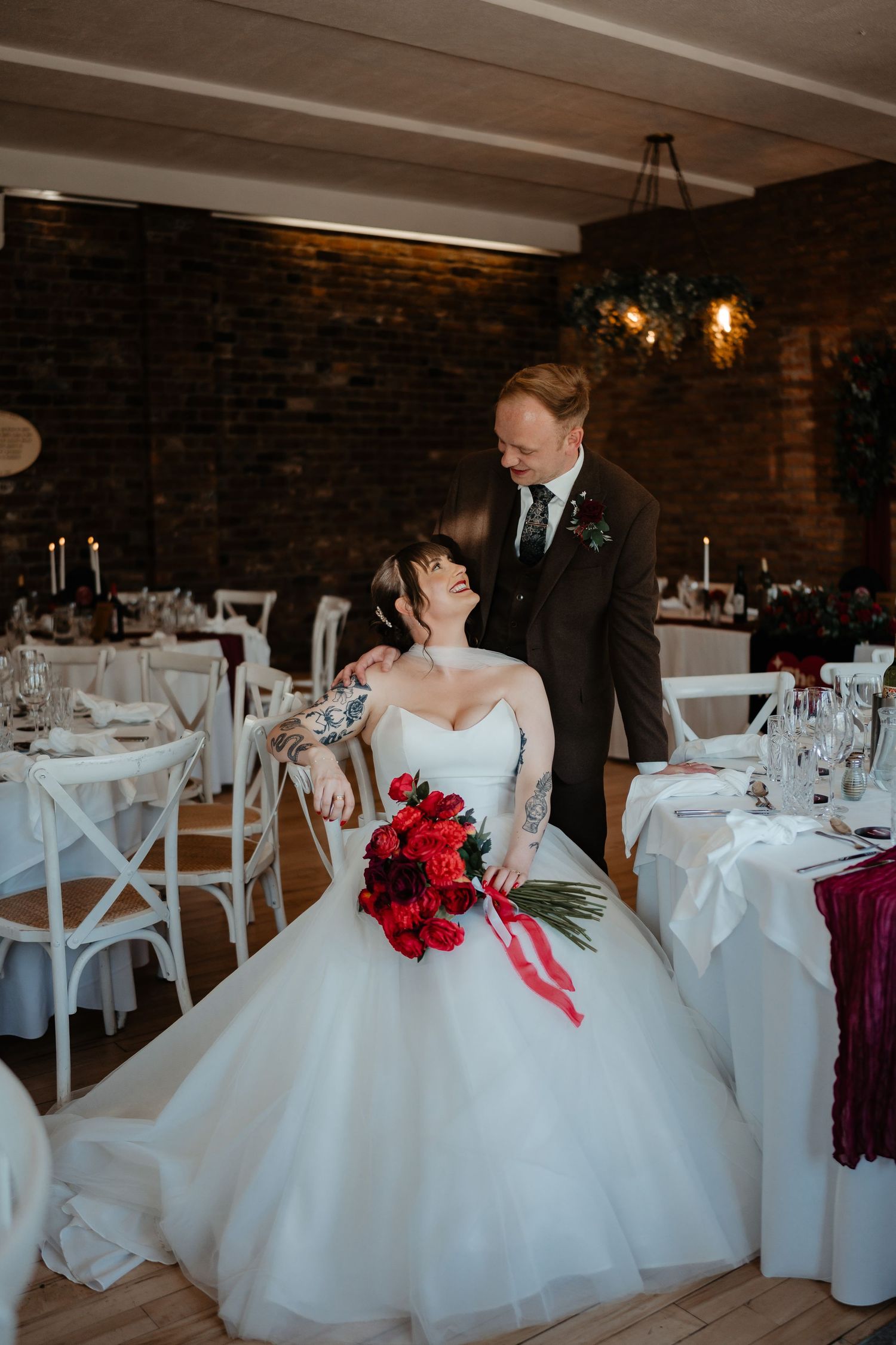 Bride in white ballgown holding red bouquet sits as groom stands beside her in decorated wedding reception venue.