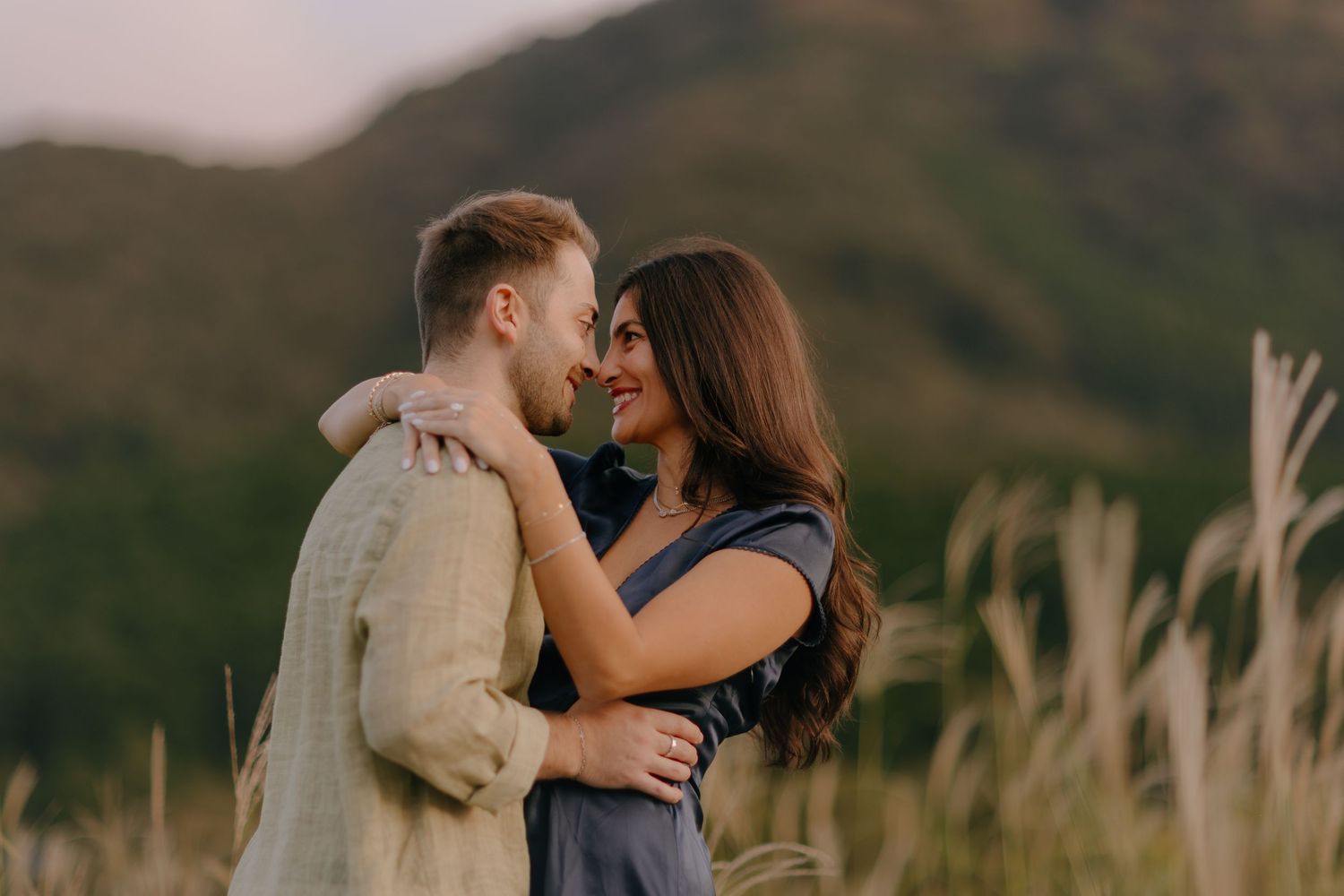 A romantic couple embraces in front of rolling hills at sunset with tall grass swaying in the foreground.