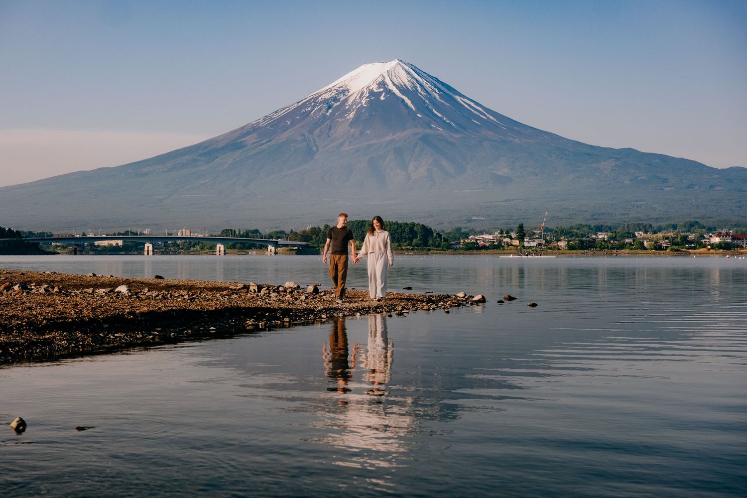 Couple standing by a serene lake with Mount Fuji snow-capped peak reflected in calm waters on a clear day.