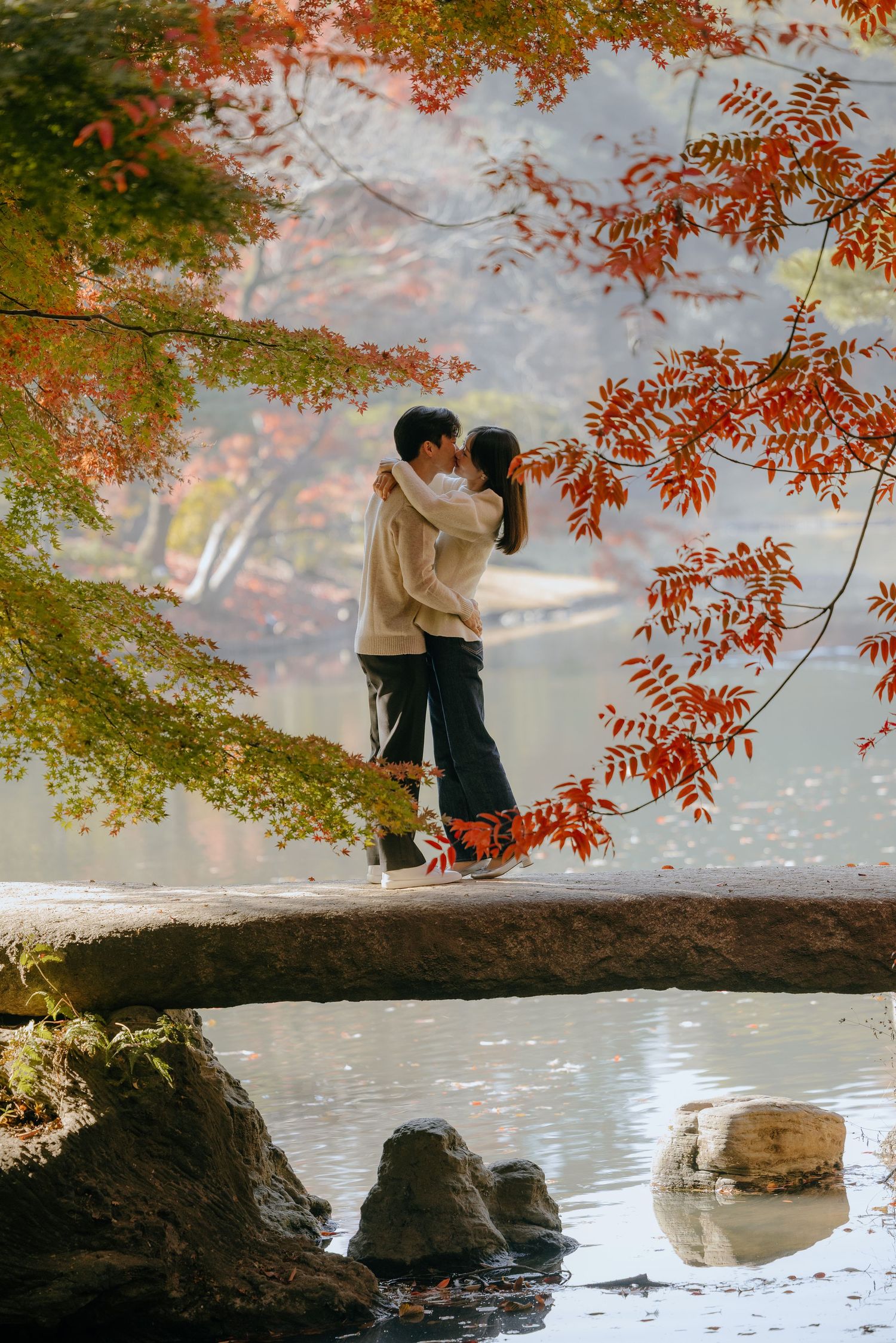 Two people share a romantic moment on a tree branch over water surrounded by vibrant autumn foliage at sunset.