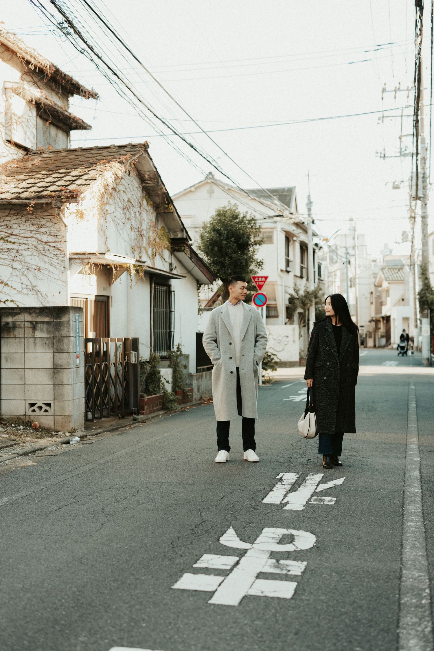 Two people in winter coats stand on a quiet residential street in Japan with road markings and power lines overhead.