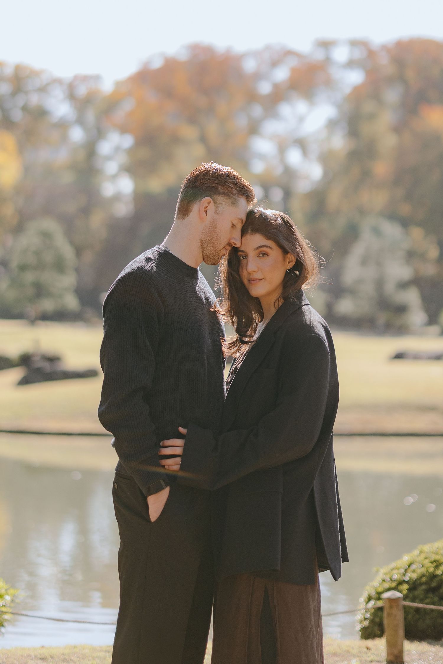 A couple in black coats shares an intimate moment by a serene lake during autumn.