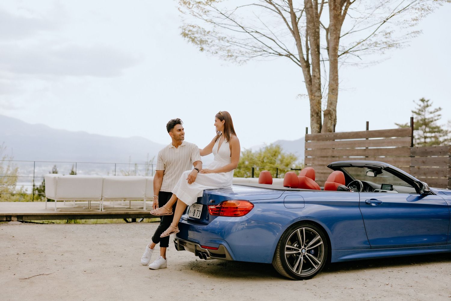 A couple sits on a blue BMW convertible parked on a scenic overlook with mountains in the background.