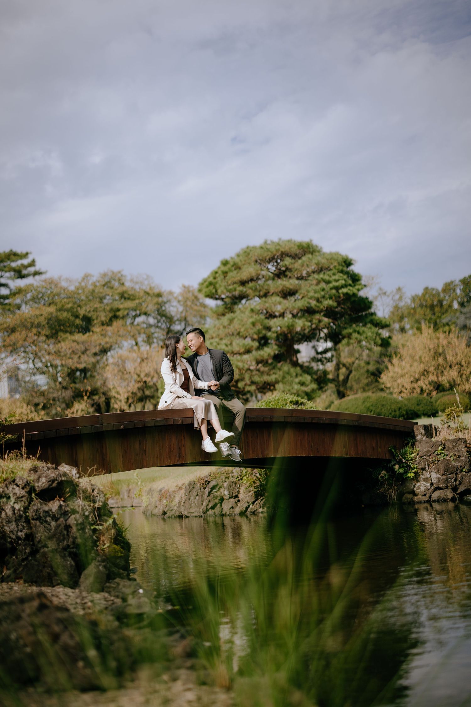 Two people sit together on a curved wooden bridge over tranquil water surrounded by autumn trees in a peaceful garden setting.