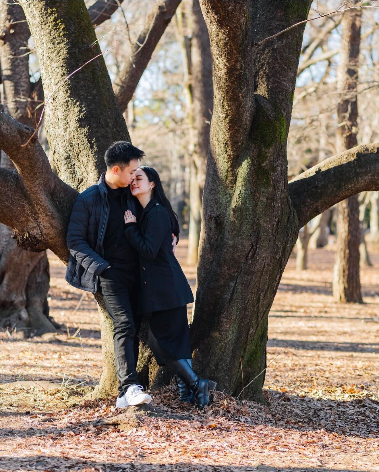 Romantic couple sharing a tender moment against a large tree trunk in an autumn park with fallen leaves on the ground.