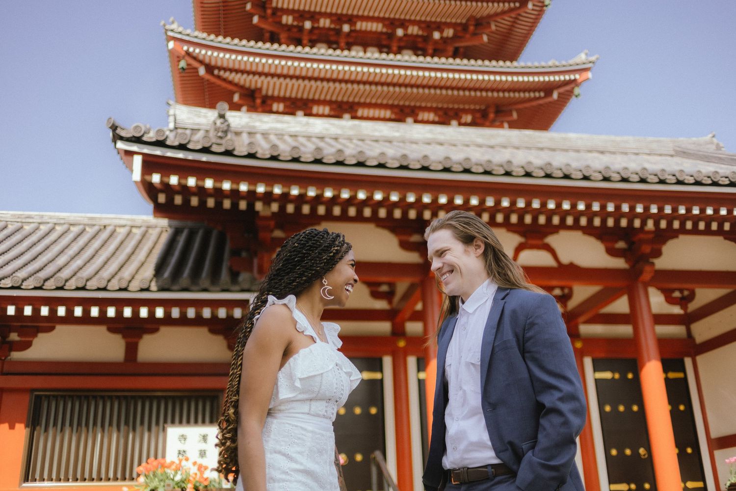 A couple poses in front of a vibrant red Japanese temple pagoda against a blue sky.