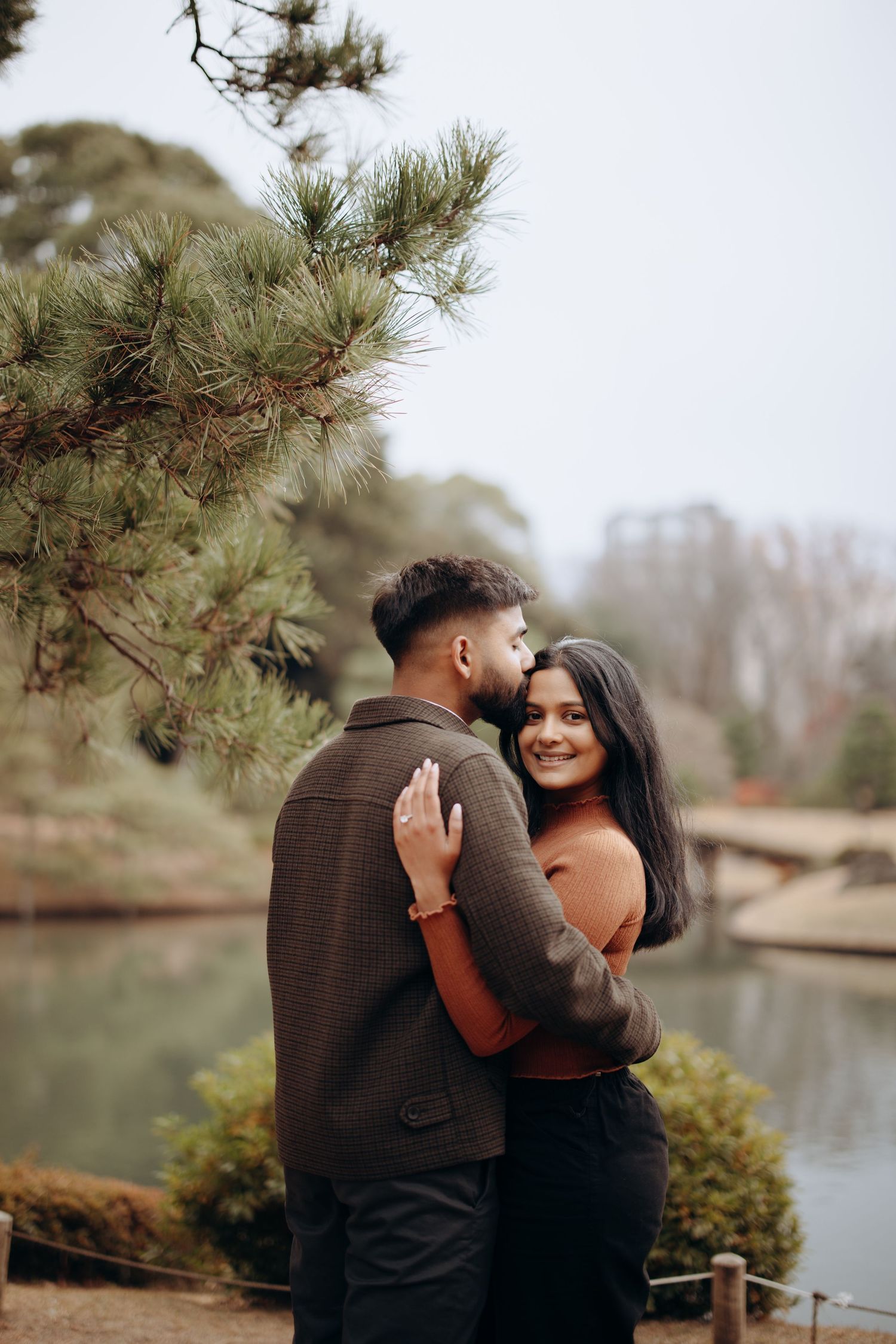 A romantic couple embrace near a lake with pine trees in a tranquil park setting on an overcast day.