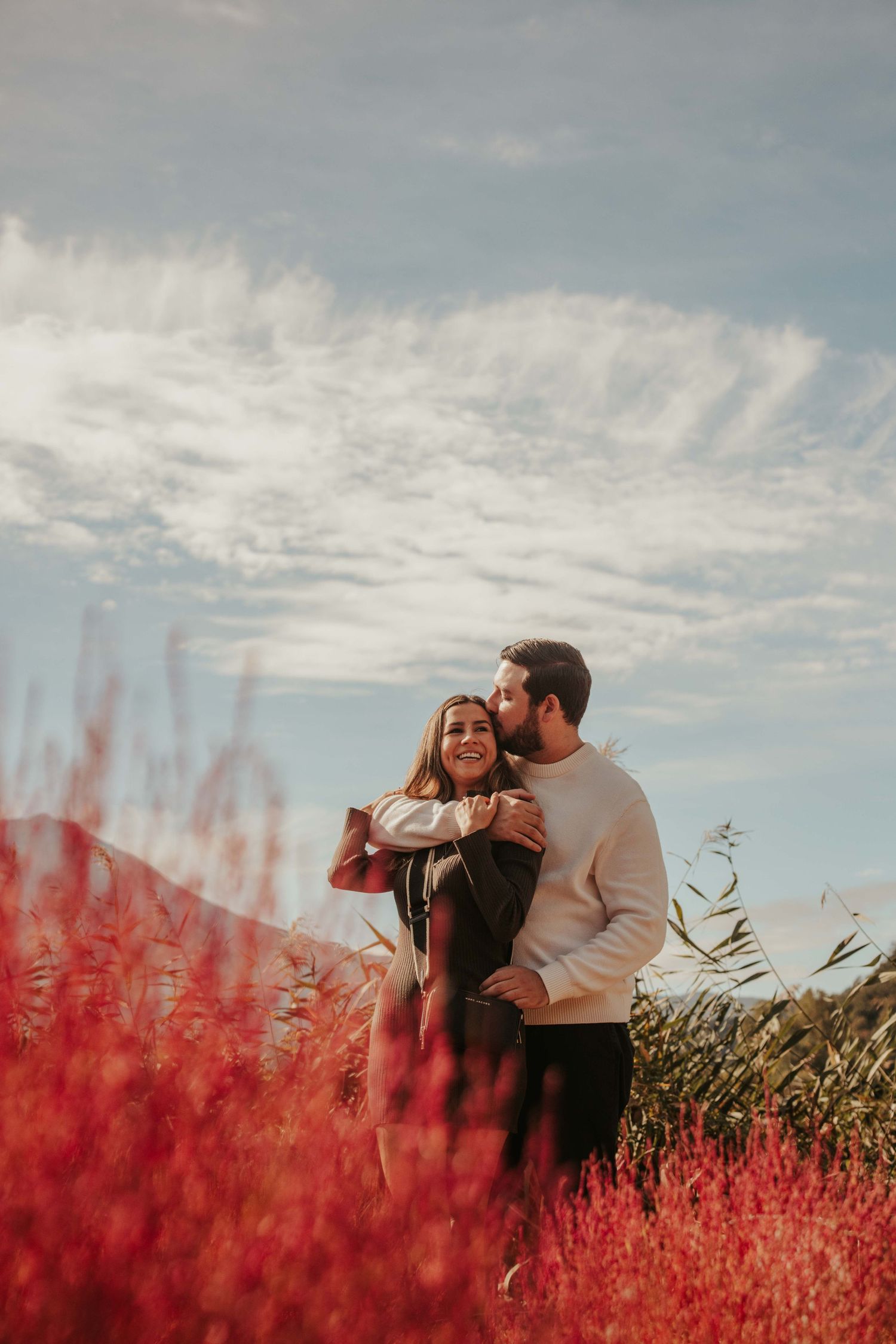 A couple shares an intimate moment among vibrant red wildflowers under dramatic clouds.
