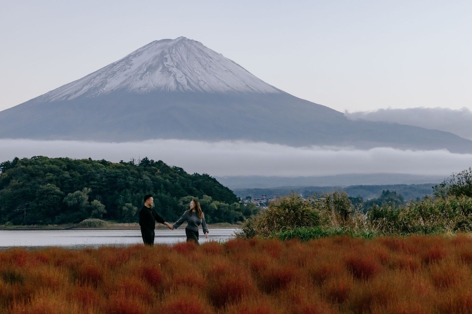 Two people stand on grassy hill overlooking Mount Fuji with misty clouds and autumn colors in the foreground.