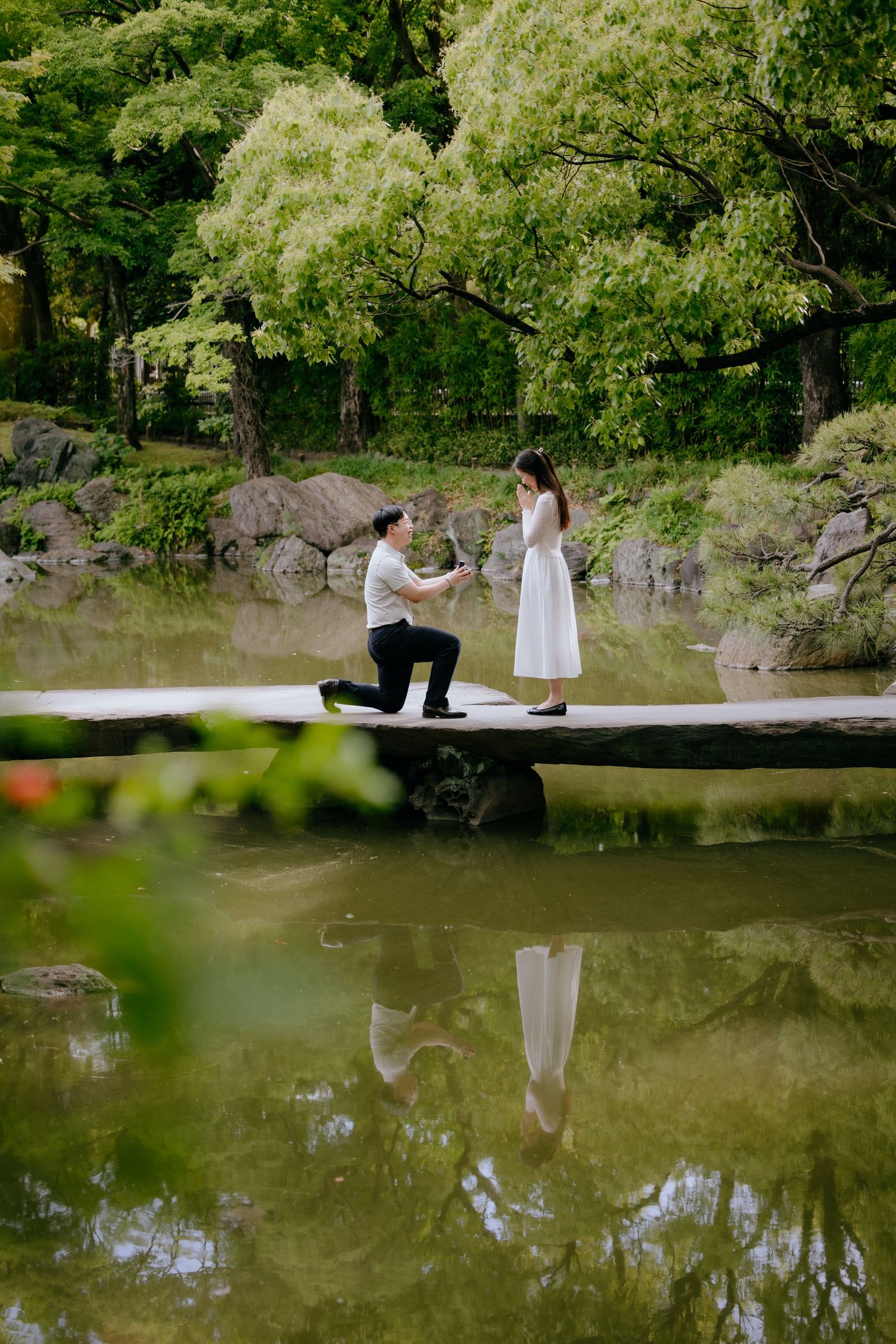 Man proposing to woman on a serene pond bridge in a lush Japanese garden, their reflection visible in the water.