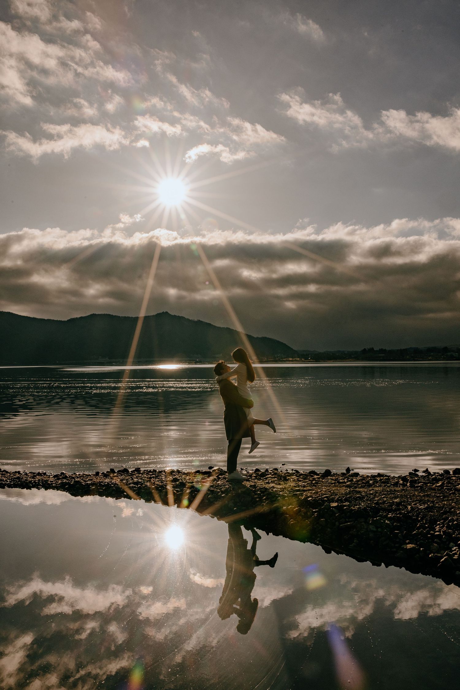 A silhouetted figure stands by a reflective lake at sunset with dramatic cloudy skies and mountain backdrop.