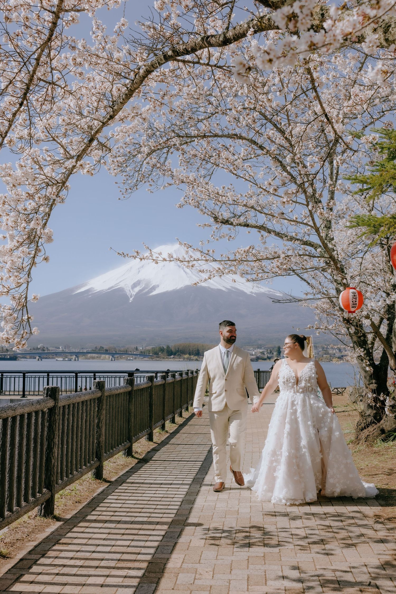 Cherry blossoms frame a scenic walkway along Mount Fuji's shore with a couple enjoying the spring scenery.