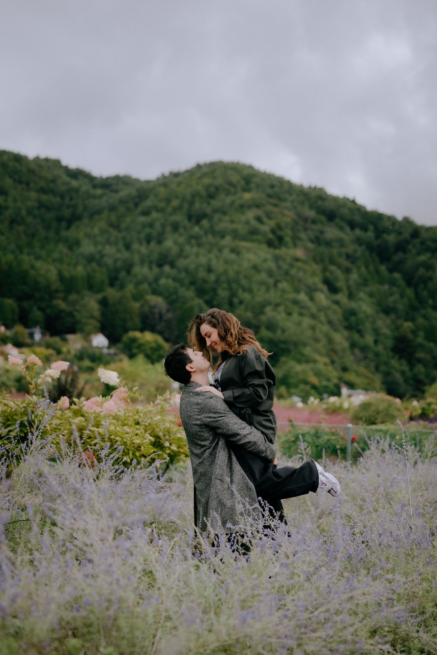 A couple embraces and spins joyfully in a field of purple wildflowers with green mountains in the background.