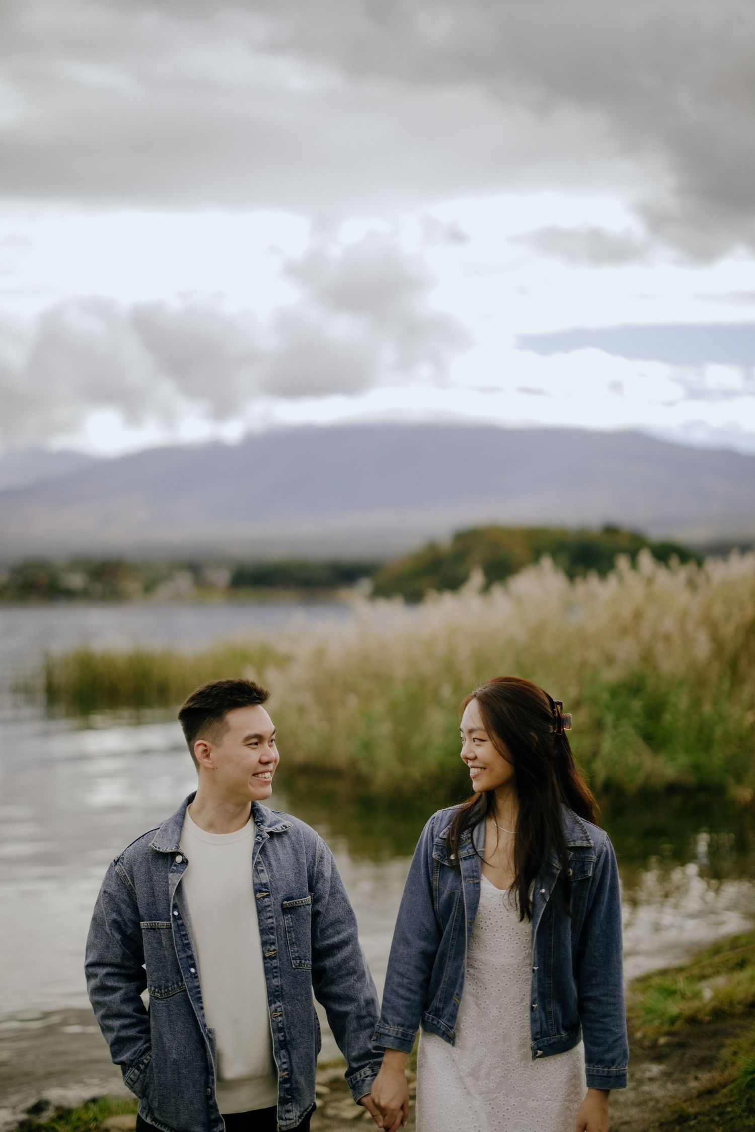 A couple in matching denim jackets holds hands while walking near a misty lake surrounded by tall grass and mountains.
