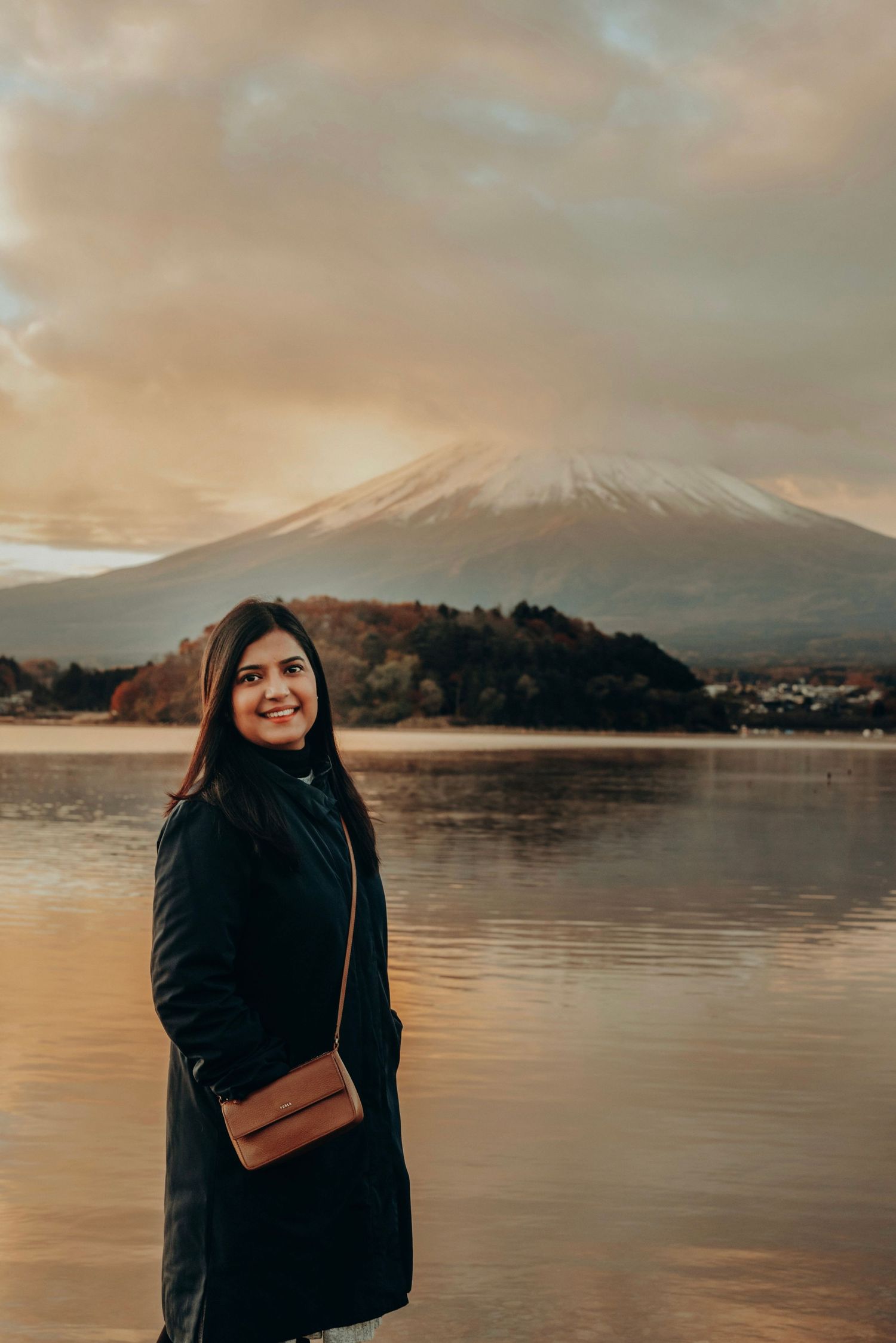 Person in black coat stands by lake with Mount Fuji visible in background during golden sunset in Japan.