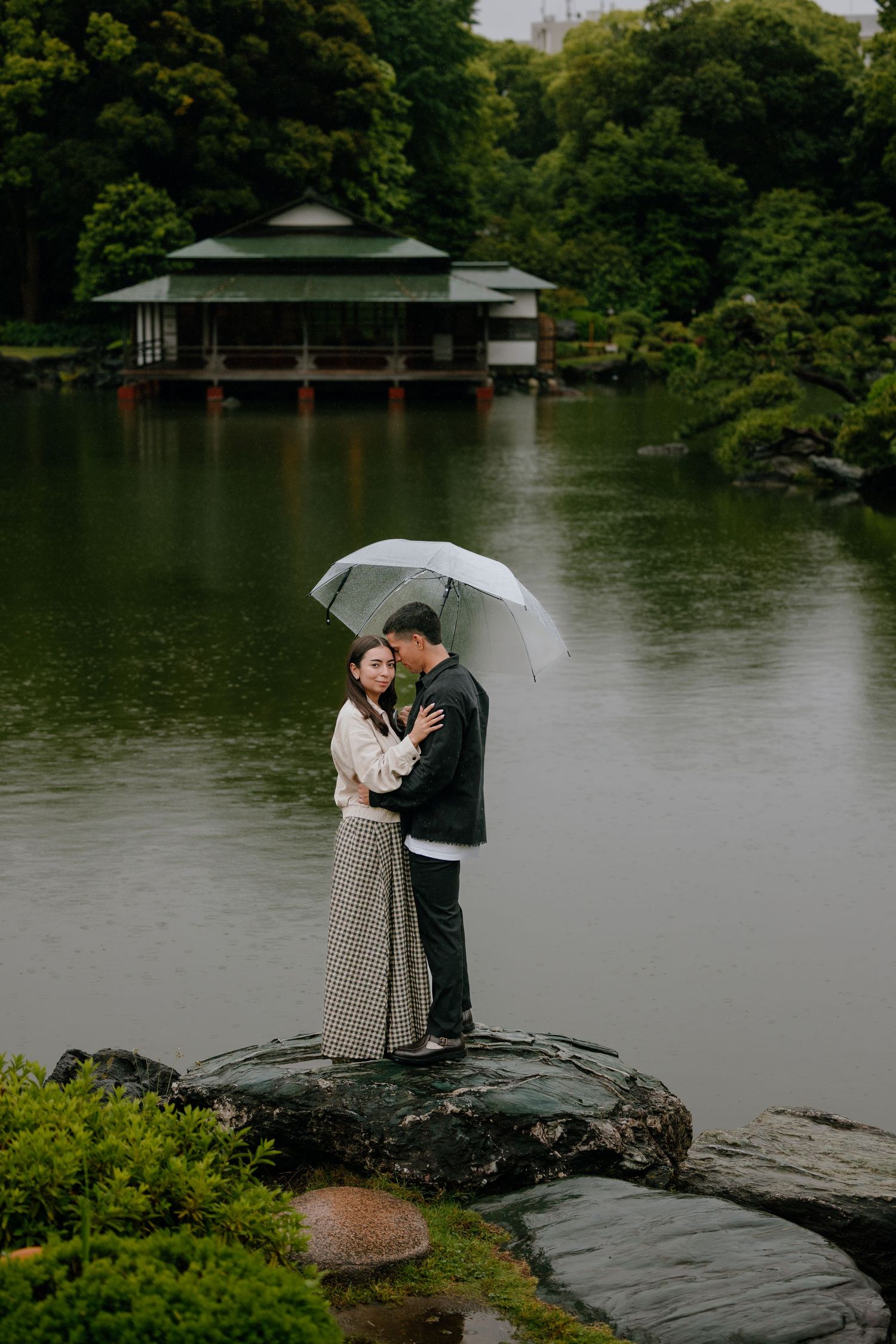 Couple embracing under white umbrella on rocks by serene Japanese garden pond with traditional teahouse backdrop.