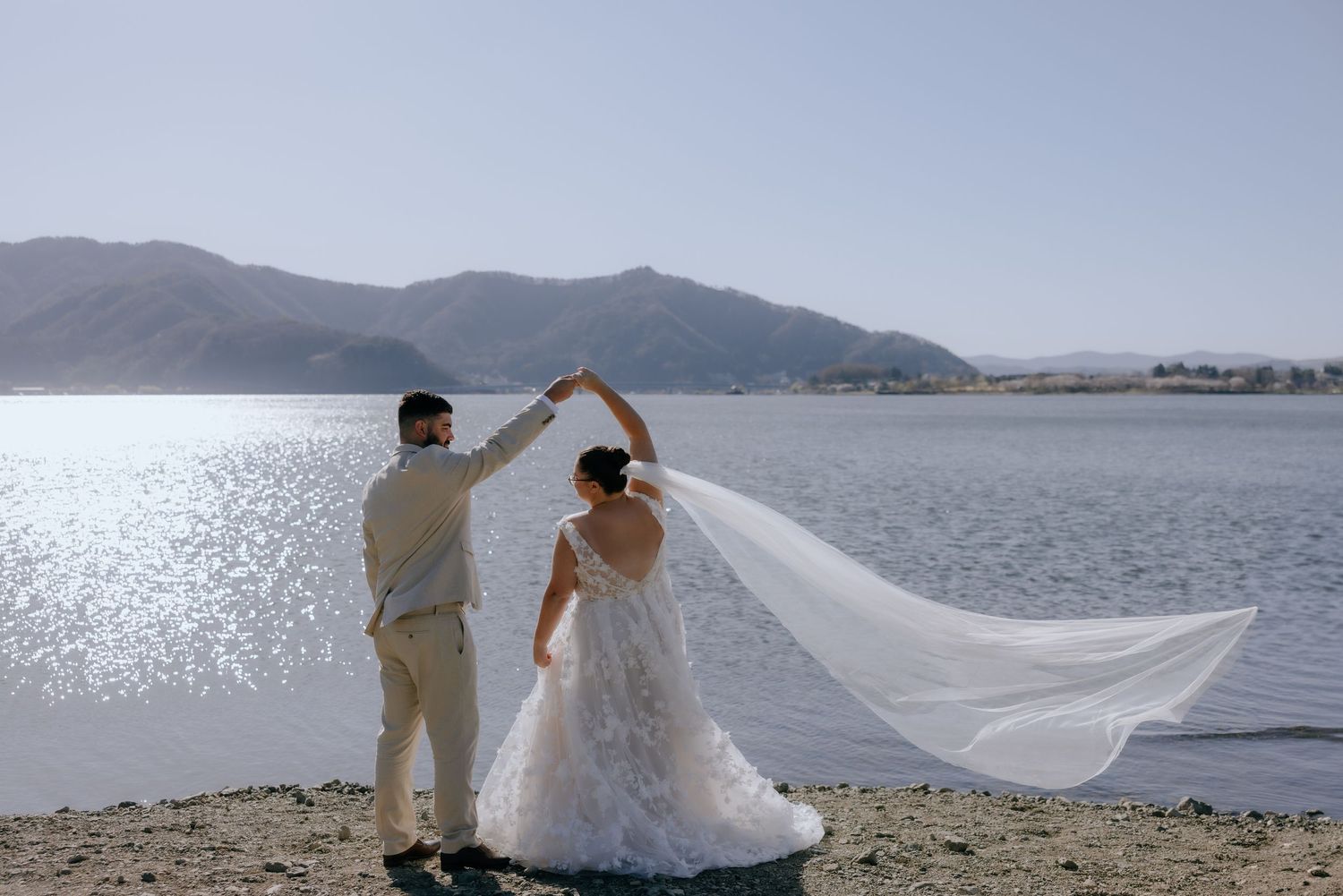 A wedding couple stands at the water's edge while a flowing veil creates a romantic silhouette against the lake view.