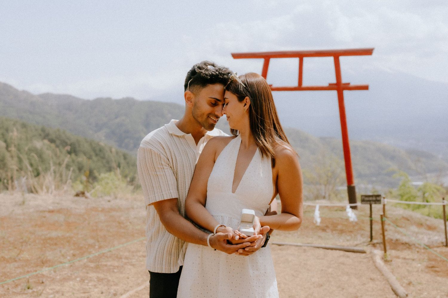 A romantic couple in white clothing stand together in front of a red torii gate with mountains in the background.