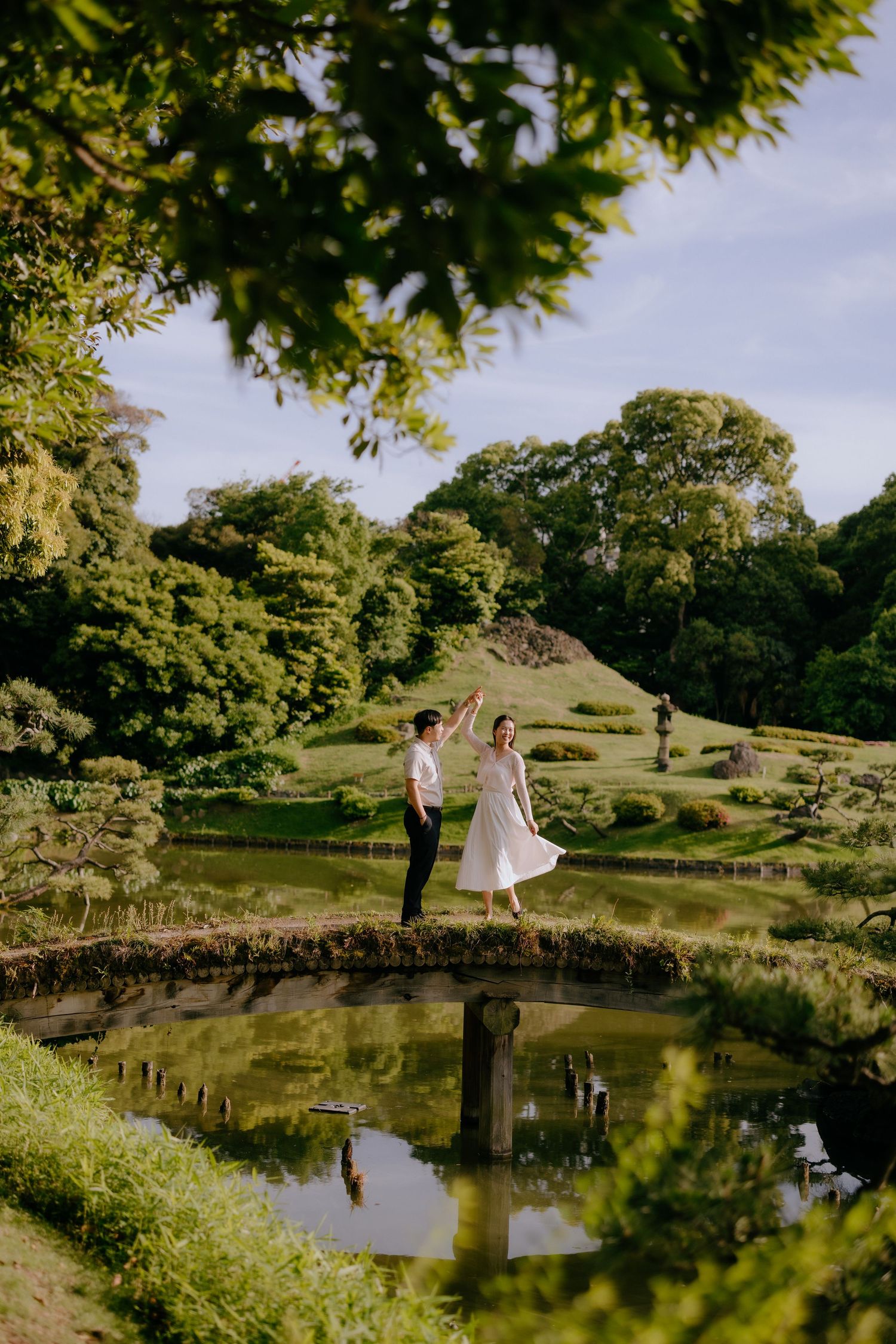 Couple dancing on a stone bridge over a serene pond in a lush Japanese garden on a sunny day.