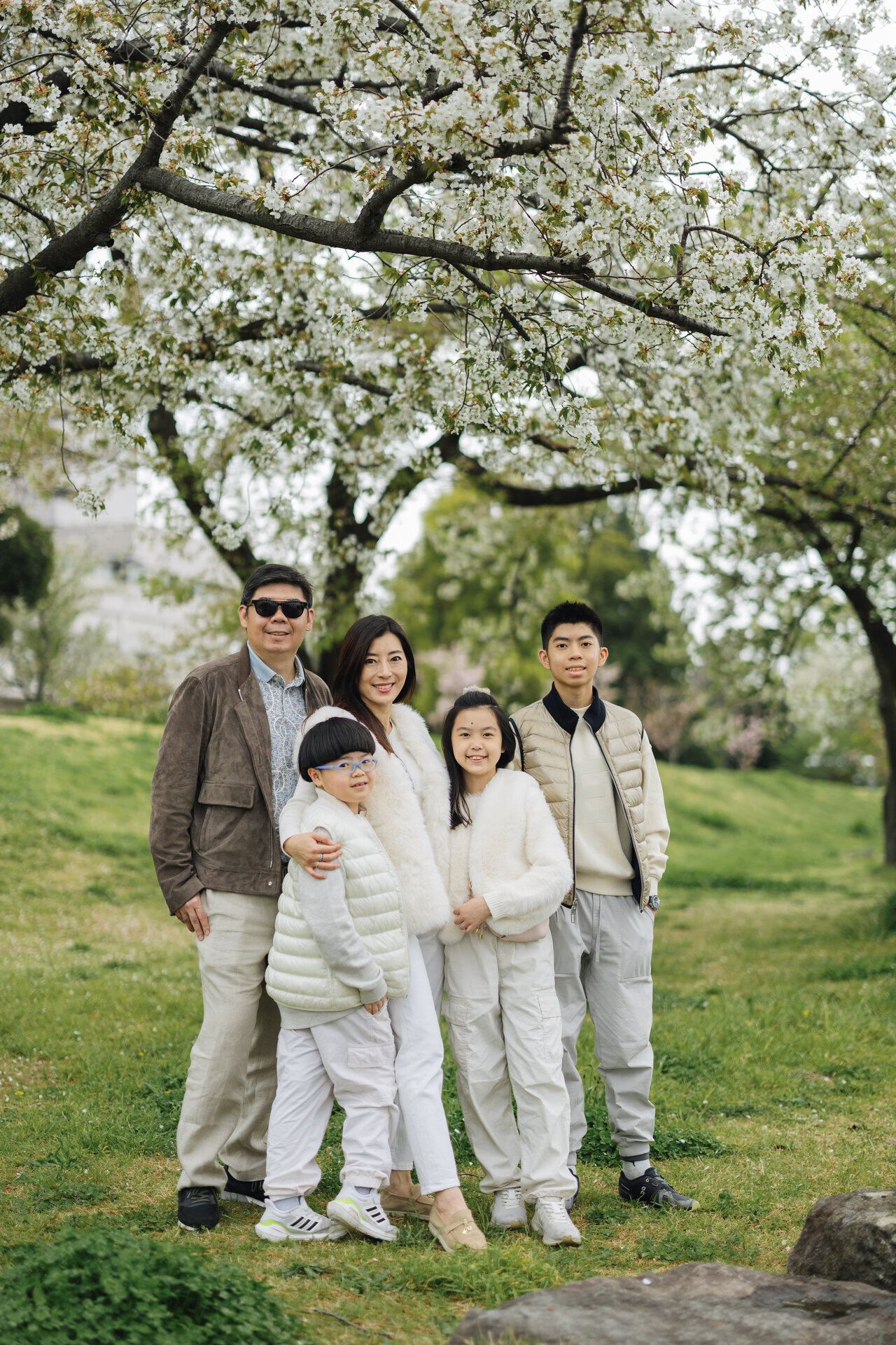 Family in coordinated white and neutral outfits poses together under blooming cherry blossom trees in a spring garden.
