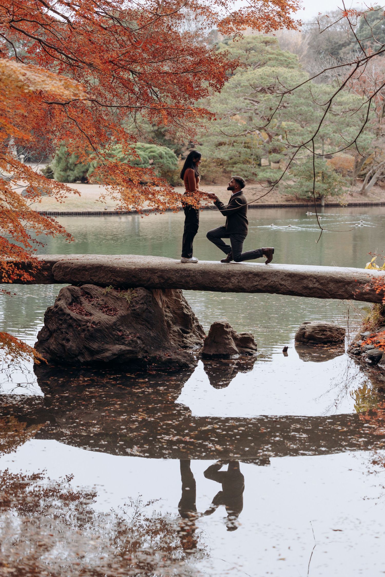 Couple's romantic marriage proposal on a stone bridge over a serene lake surrounded by vibrant autumn foliage.