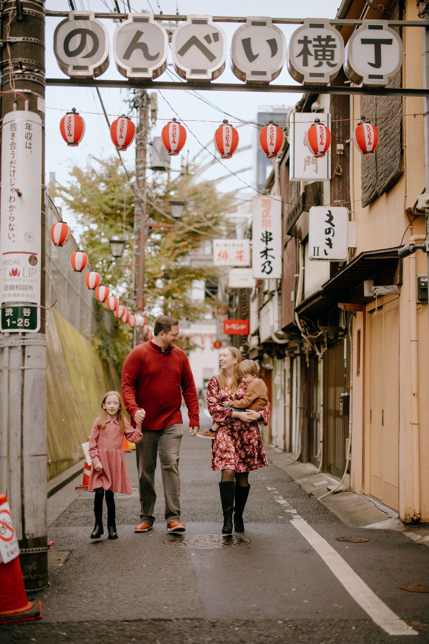 Family walking down a traditional Japanese street decorated with red lanterns under a 'Nonbei' archway in winter.