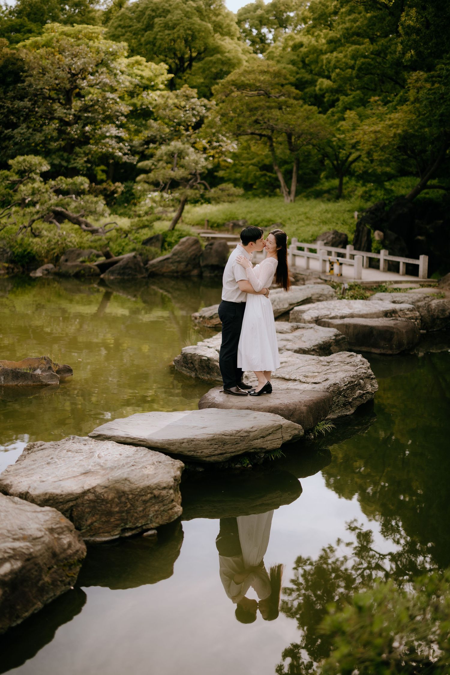 Couple kissing on stepping stones over a serene pond in a lush Japanese garden.