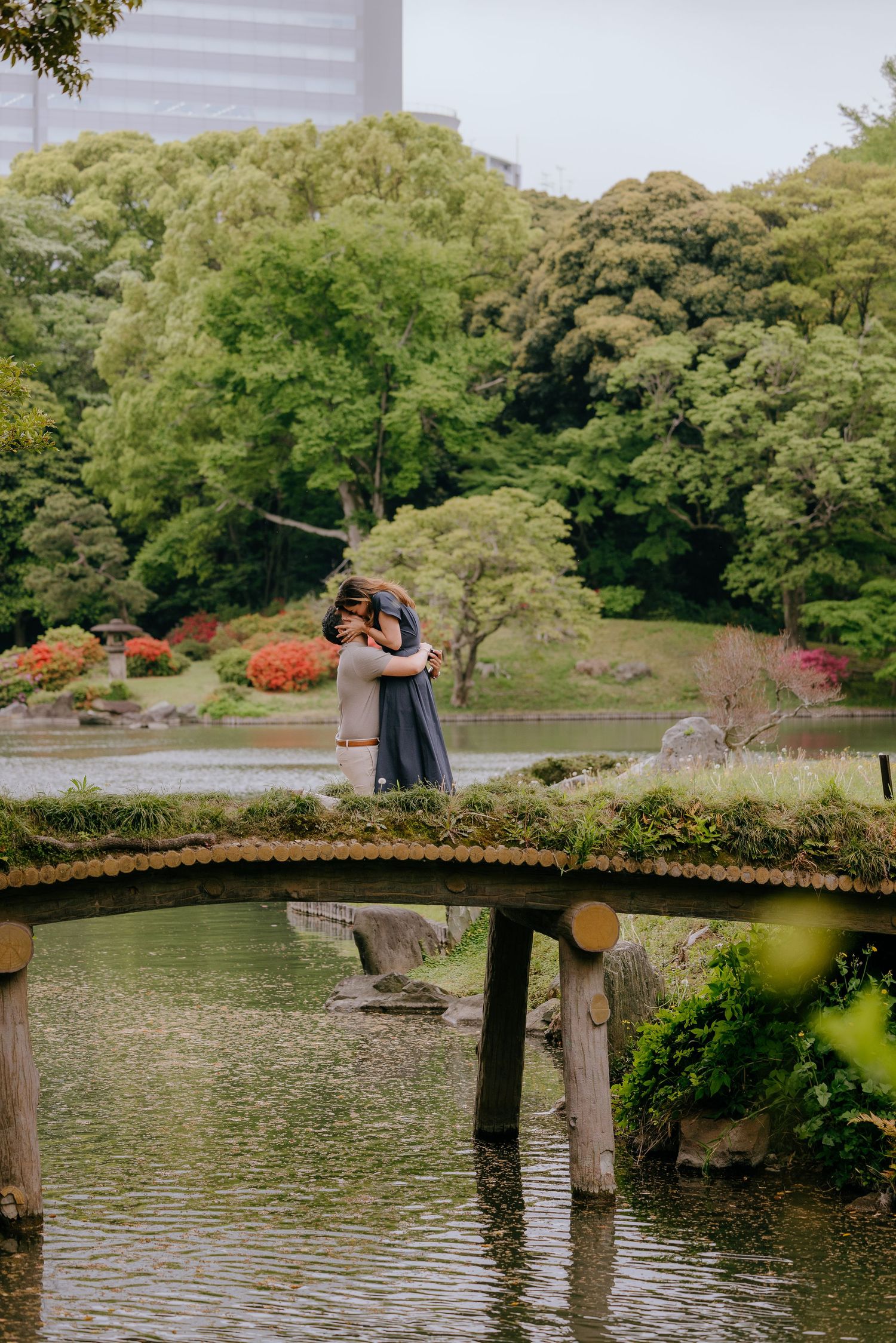 Couple embracing on a wooden bridge in a lush Japanese garden with a tranquil pond and vibrant greenery.