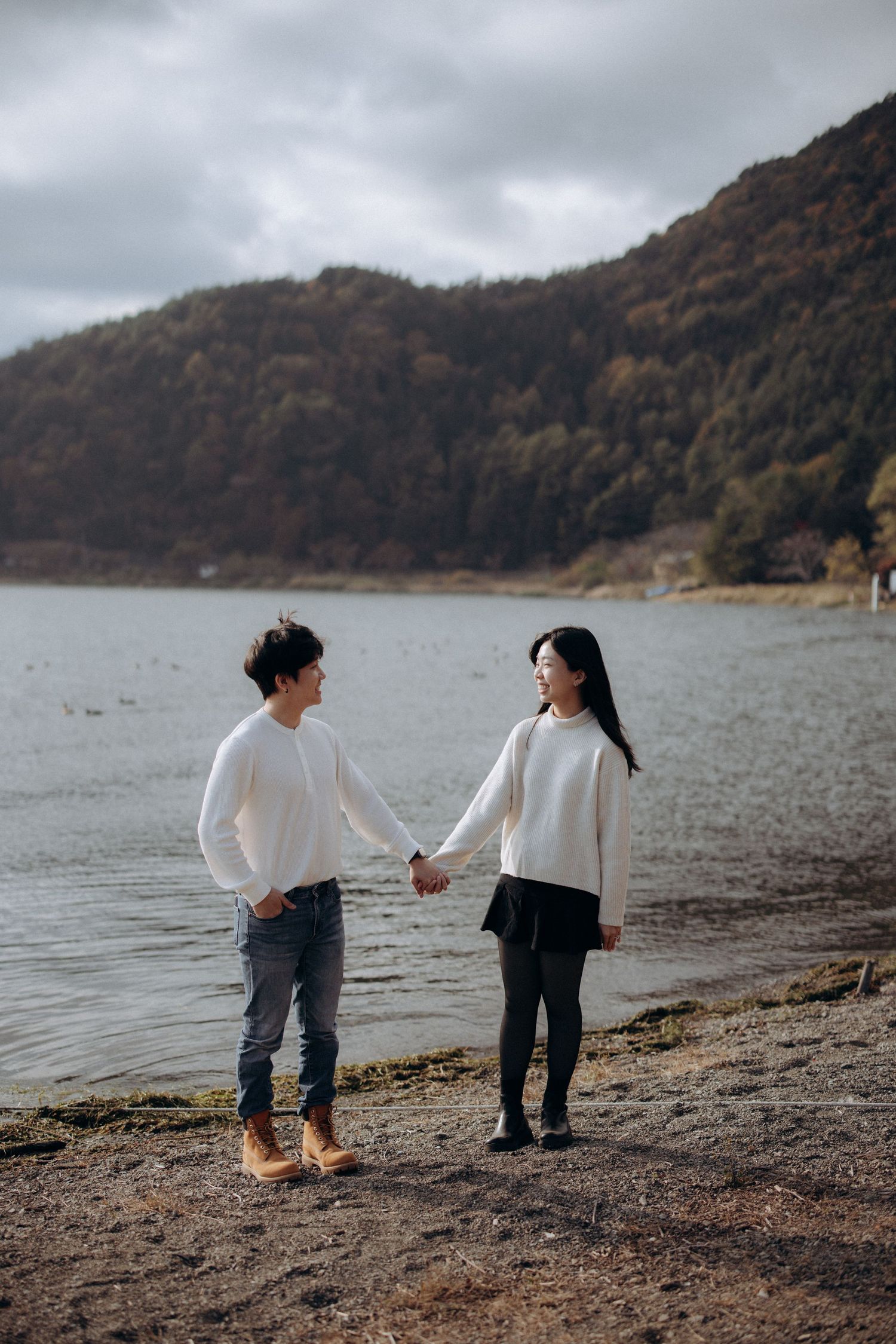 Two people in white sweaters hold hands while standing on a shoreline with moody mountains in the background.