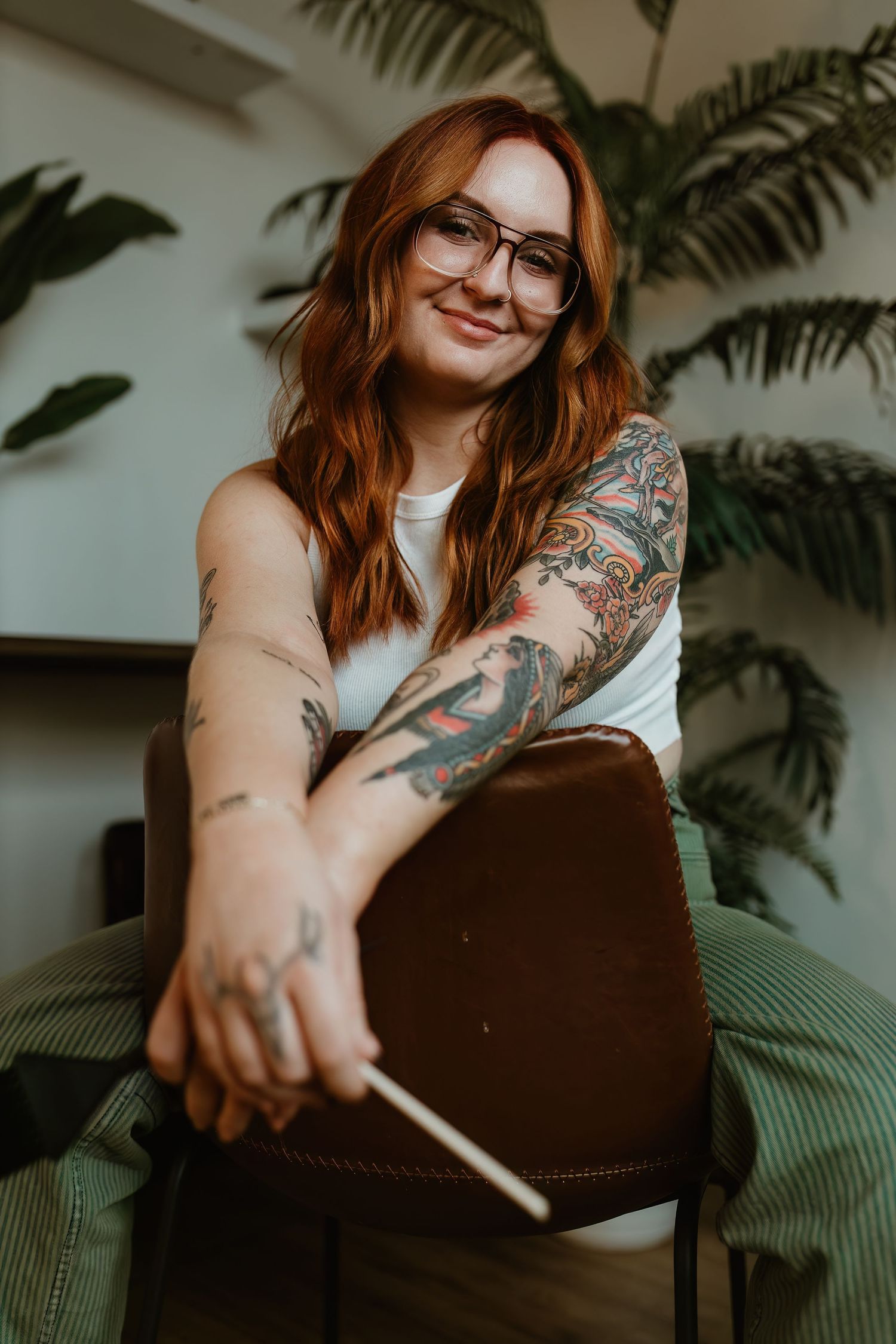 A person with red hair and tattoos sits casually on a chair with indoor plants in the background.