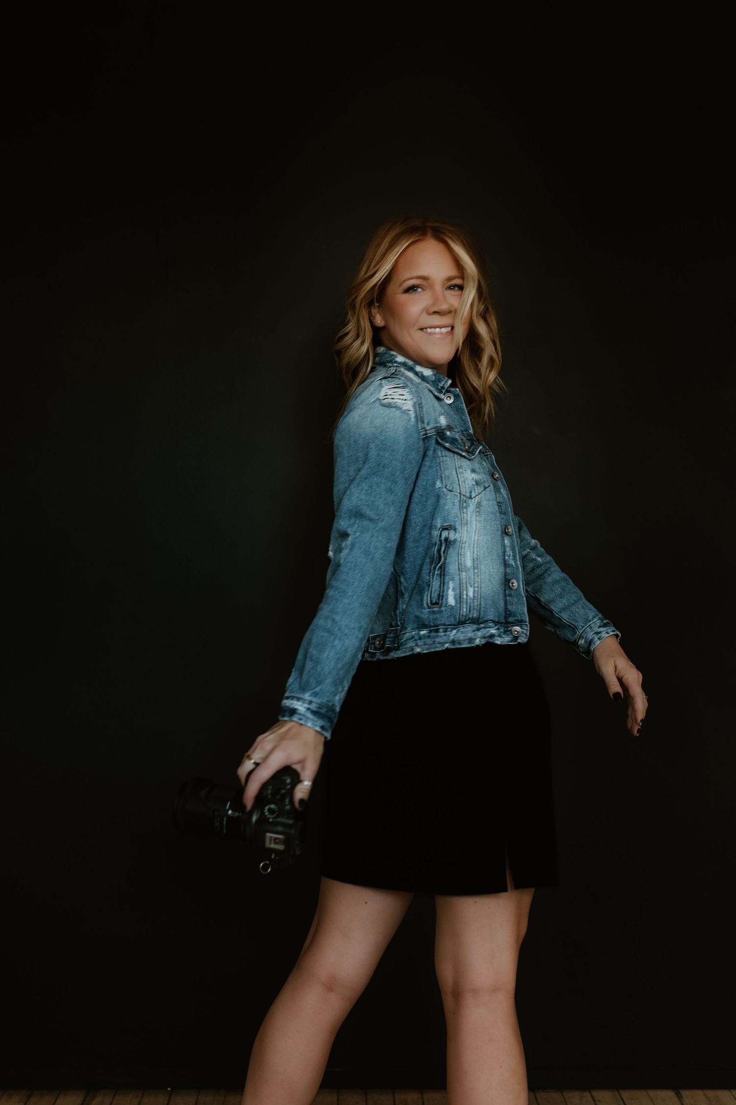 Dynamic portrait shot of photographer in denim jacket and black skirt against dark backdrop.