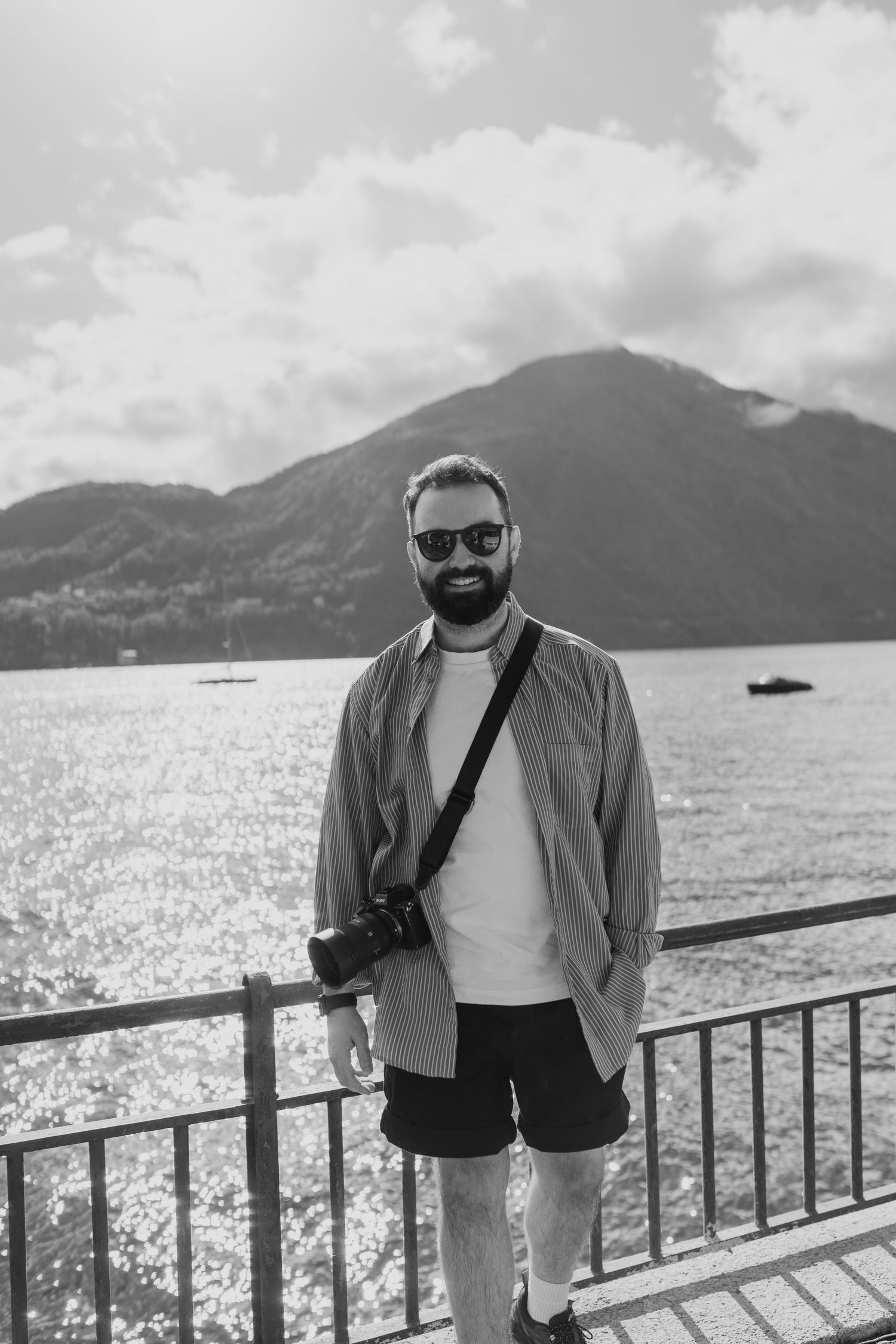 Man standing smiling at the camera with Lake Como behind him, hes wearing a shirt and a camera is over his shoulder.