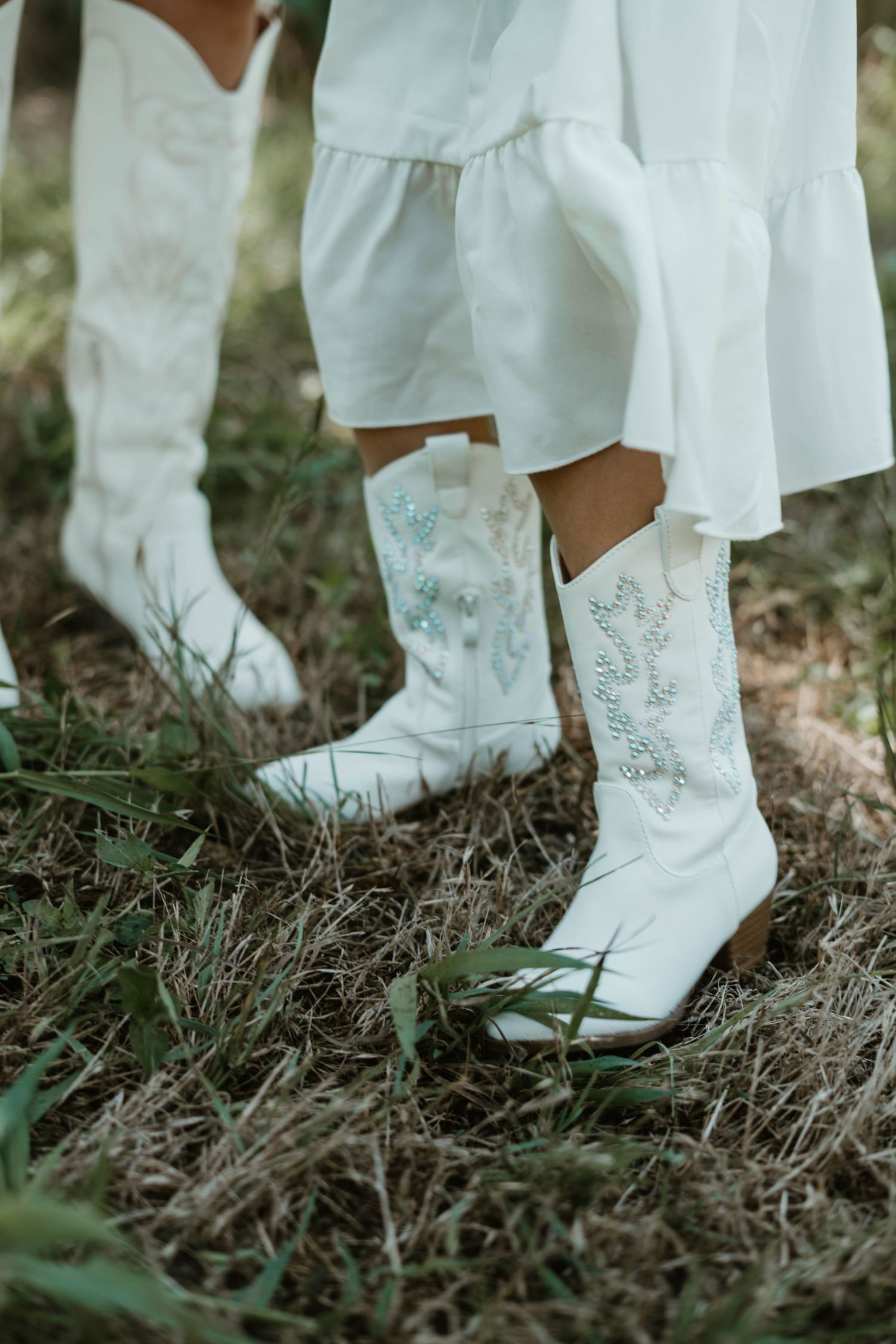 White leather cowgirl boots with floral embroidery shown on grass against a flowing white dress.