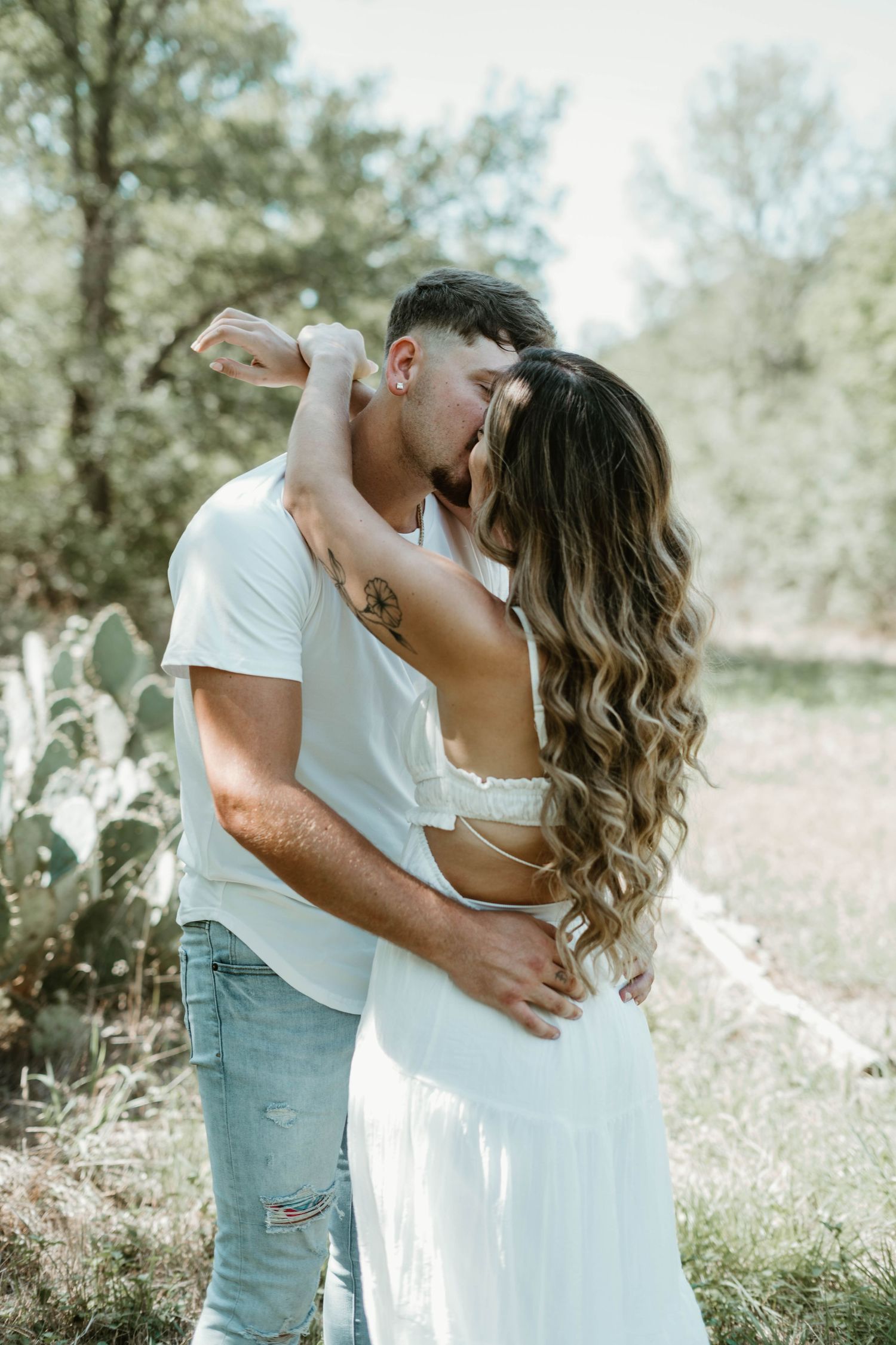 A romantic couple embraces in a sun-dappled outdoor setting with cactus plants and trees in the background.