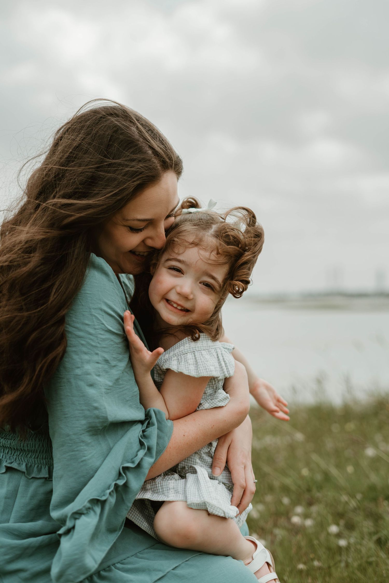 A tender moment between a parent and child embracing outdoors on a cloudy day by the water.