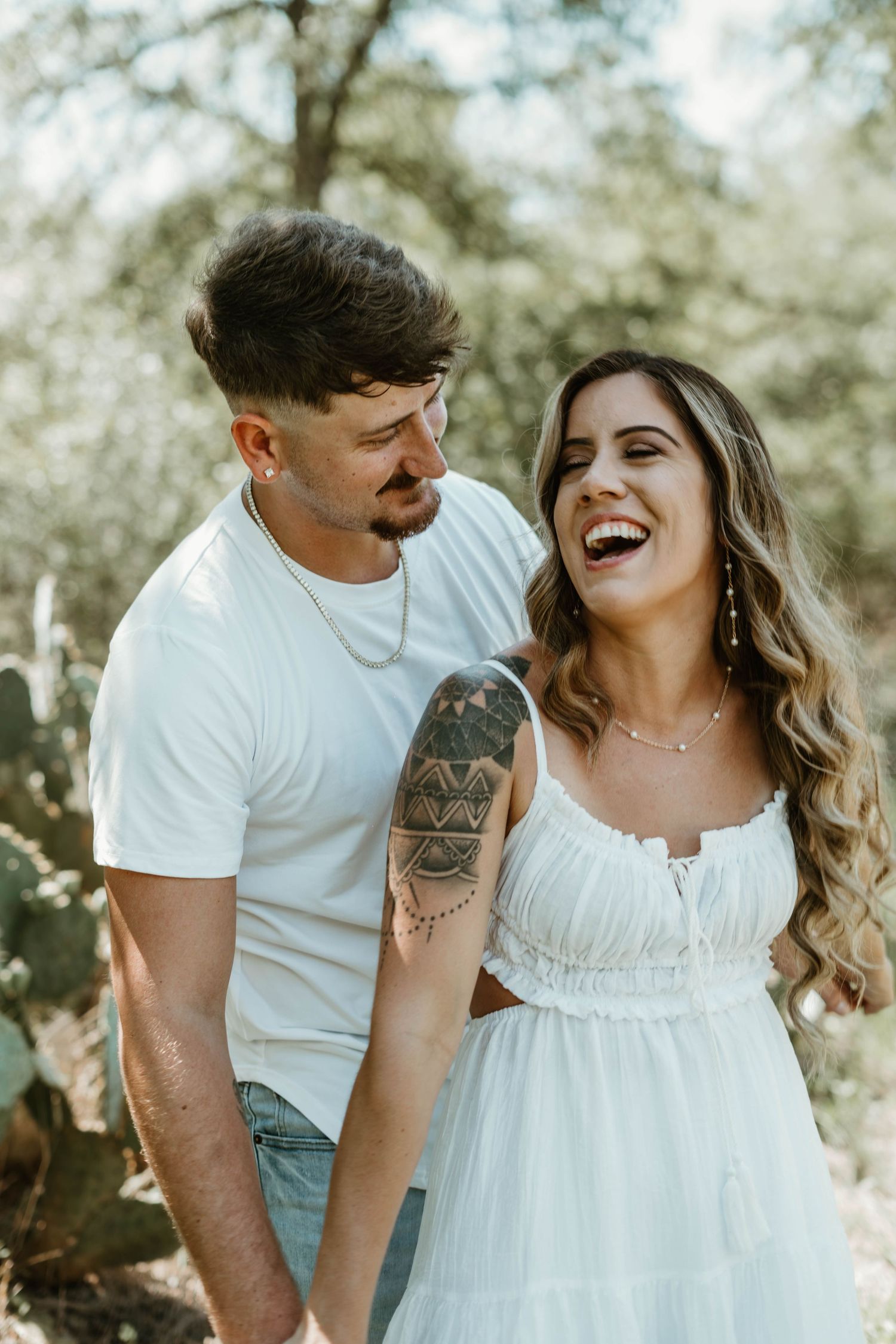 A joyful couple wearing white shares a candid moment outdoors in nature, with sunlight filtering through trees.